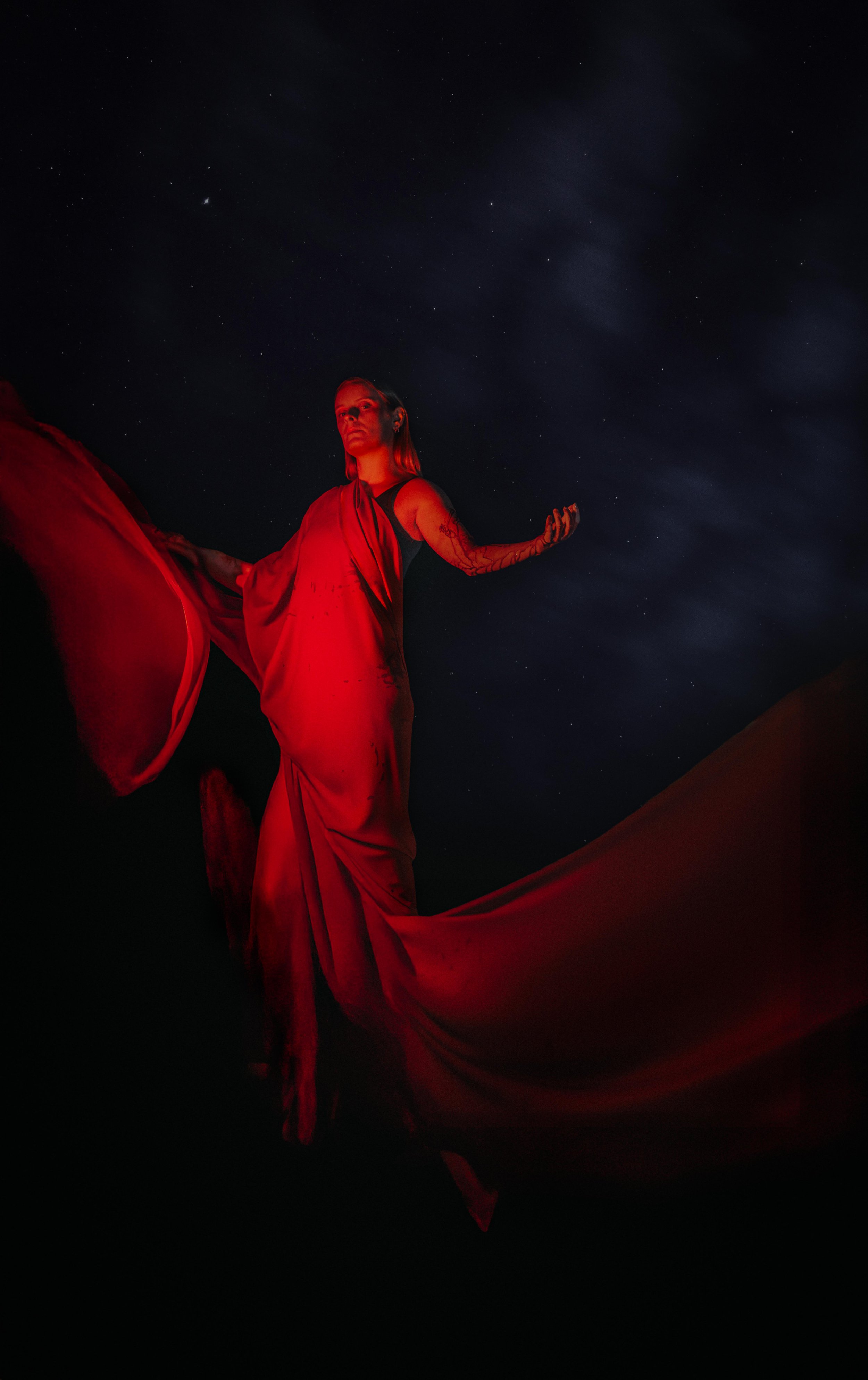A woman dressed in a flowing red garment stands against a dark night sky filled with stars, with clouds faintly visible.