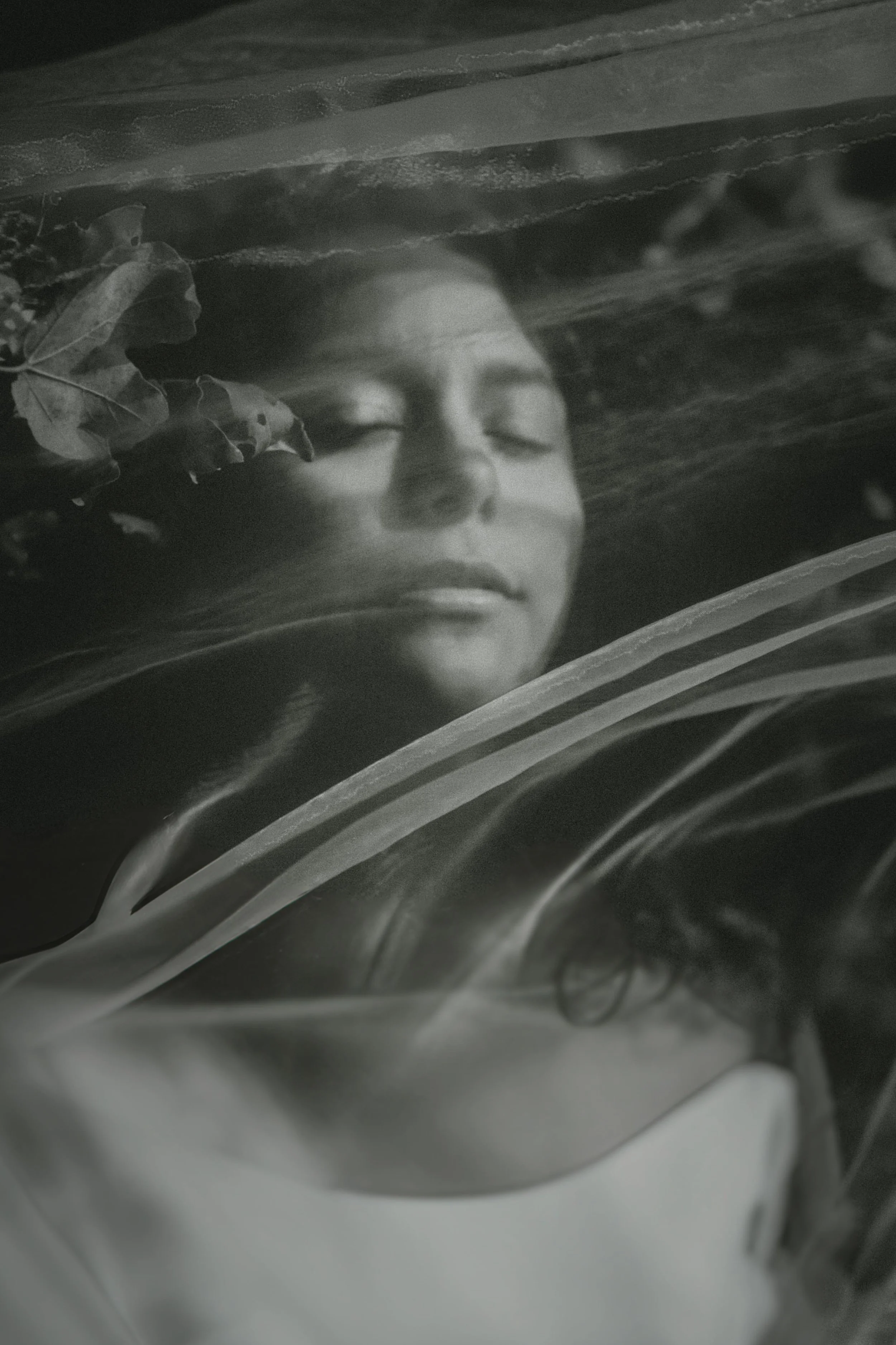 A black and white photo of a person with closed eyes, resting their head on a surface covered with feathers, leaves, and other natural elements.