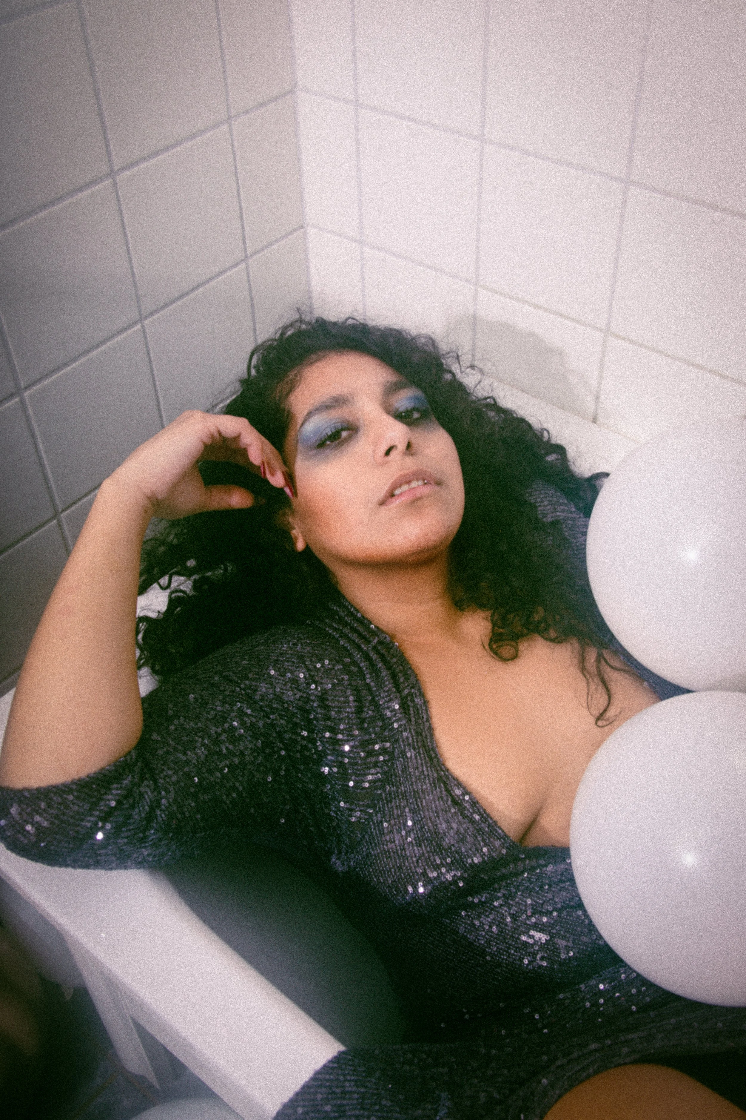 A woman with curly dark hair lying in a white tub or sink, wearing a sparkly purple top with makeup and red nail polish.