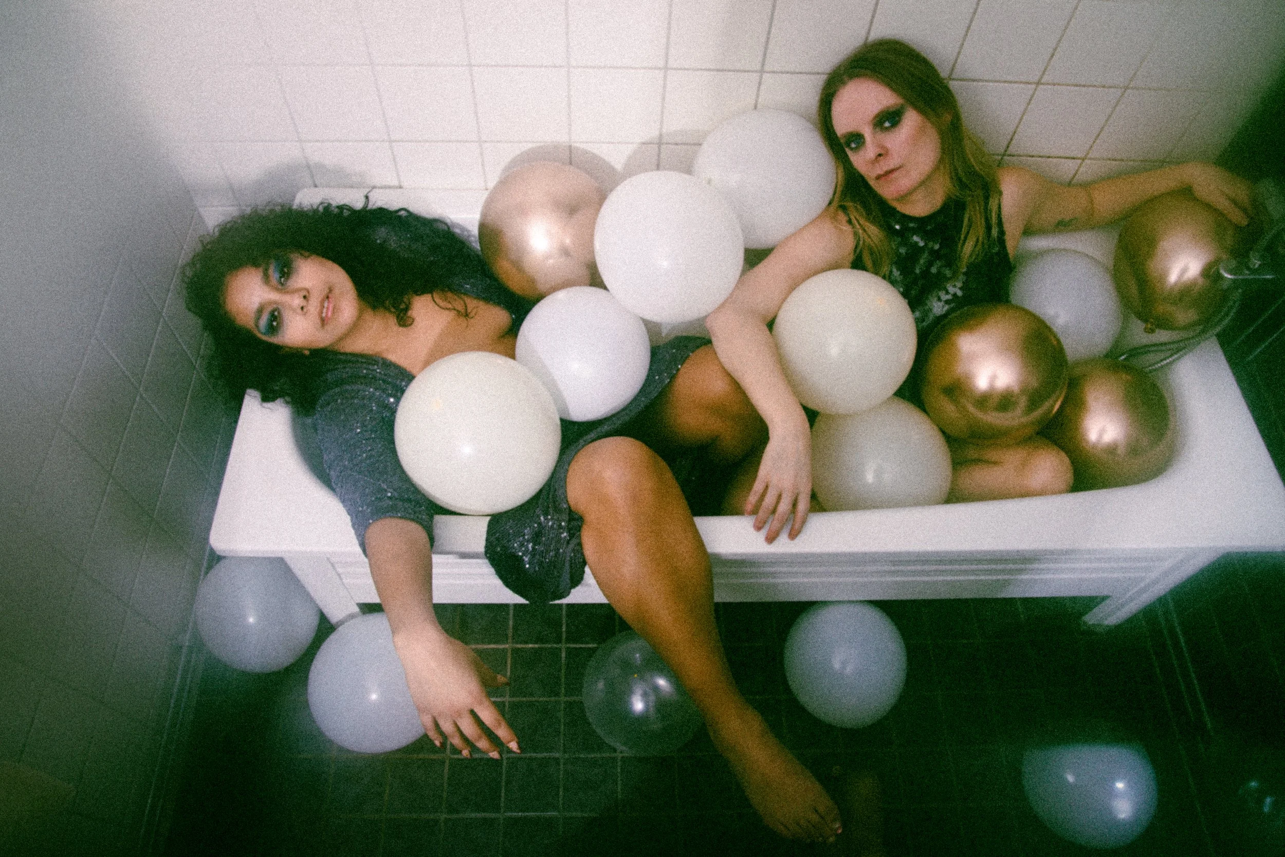 Two women are lying in a bathtub filled with white and gold balloons, with a white tiled wall in the background.