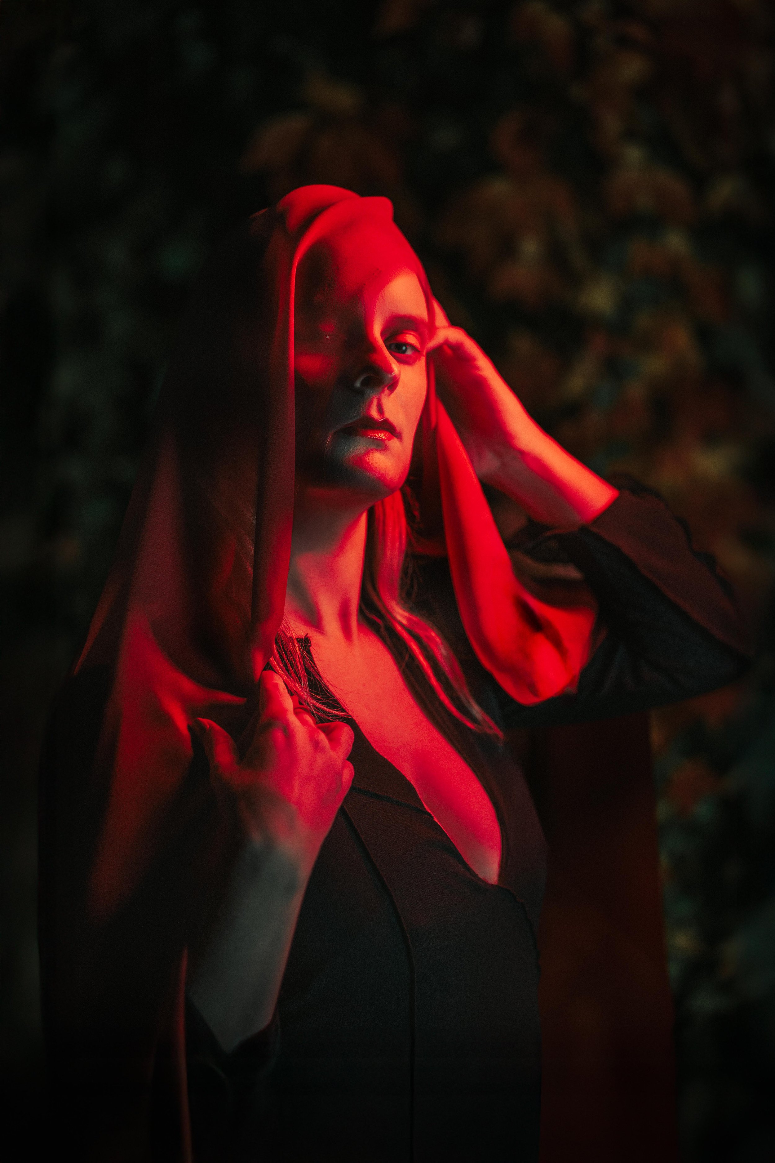 A woman with blonde hair in a bun wearing a red hooded garment, outdoors at night, illuminated by red lighting, with dark, blurry background.