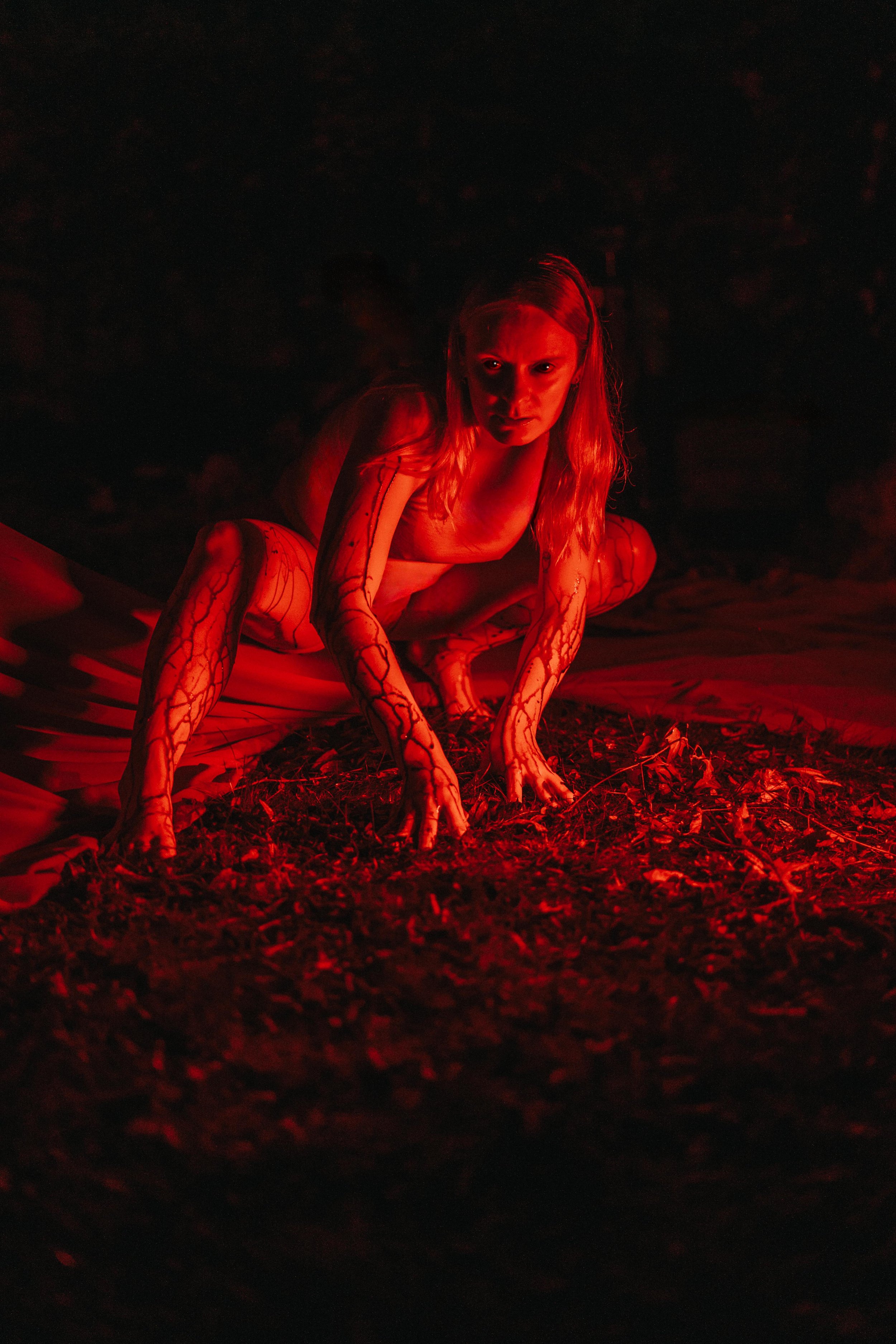 A woman crouches on the ground illuminated by red light, with intense expression, dark background, and her body covered in intricate markings or tattoos.