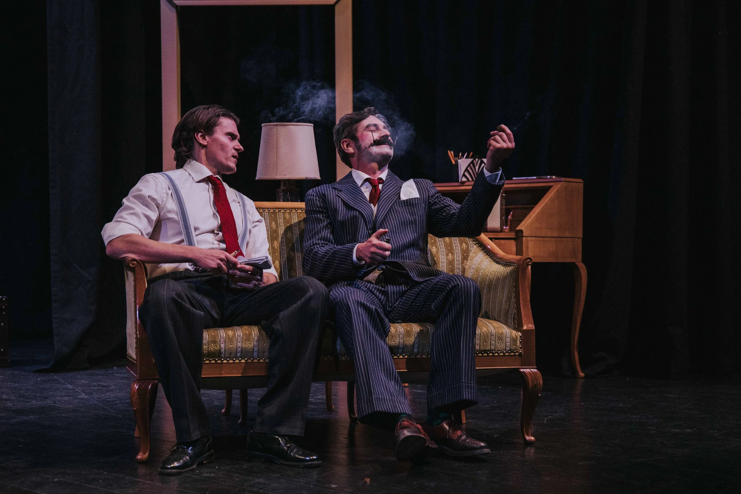 Two men in vintage attire sitting on a couch, one smoking a cigar, in a theatrical stage setting.
