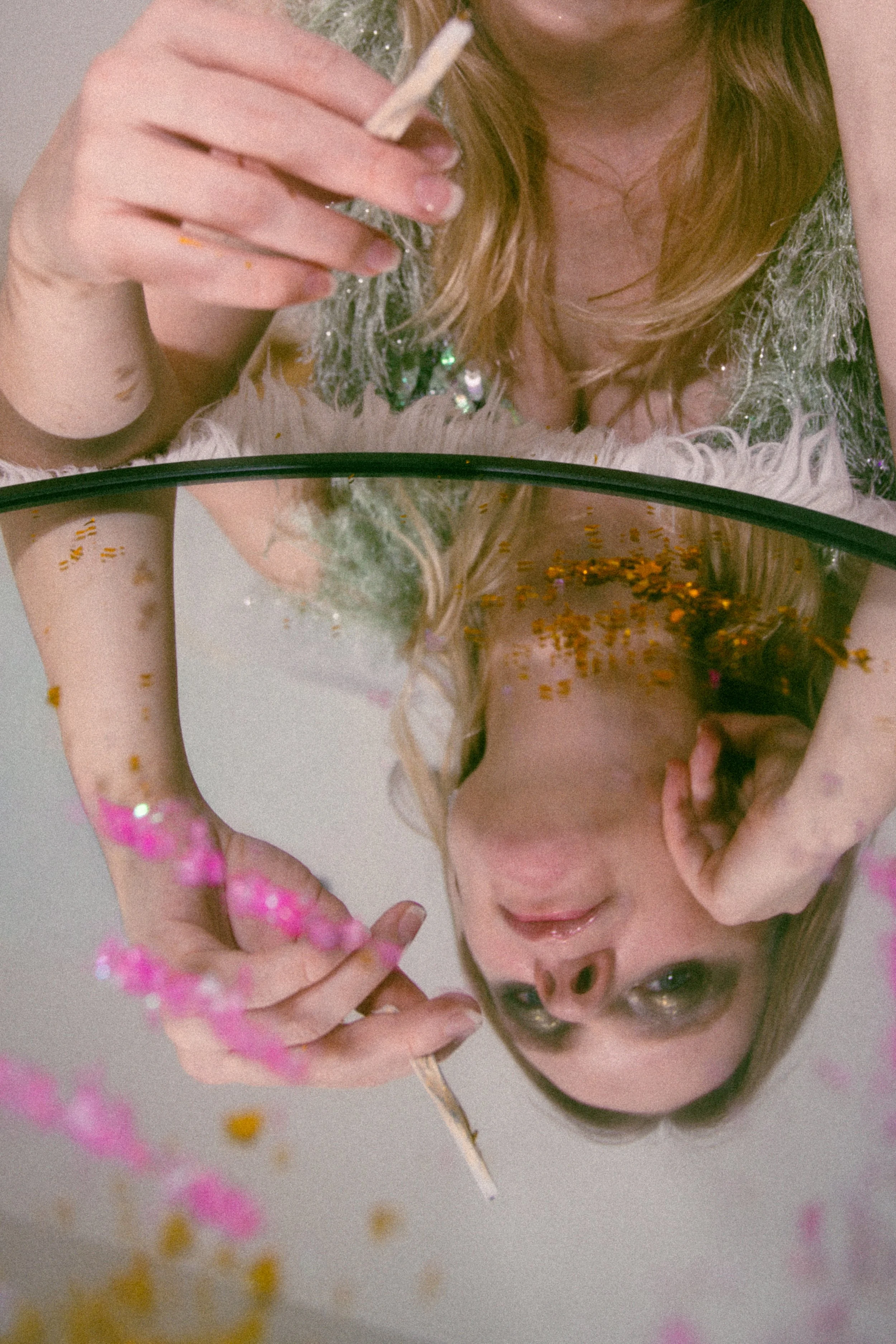 A woman looking into a mirror, holding a cigarette, with colorful glitter on her face and dress, and pink beads around her hand.