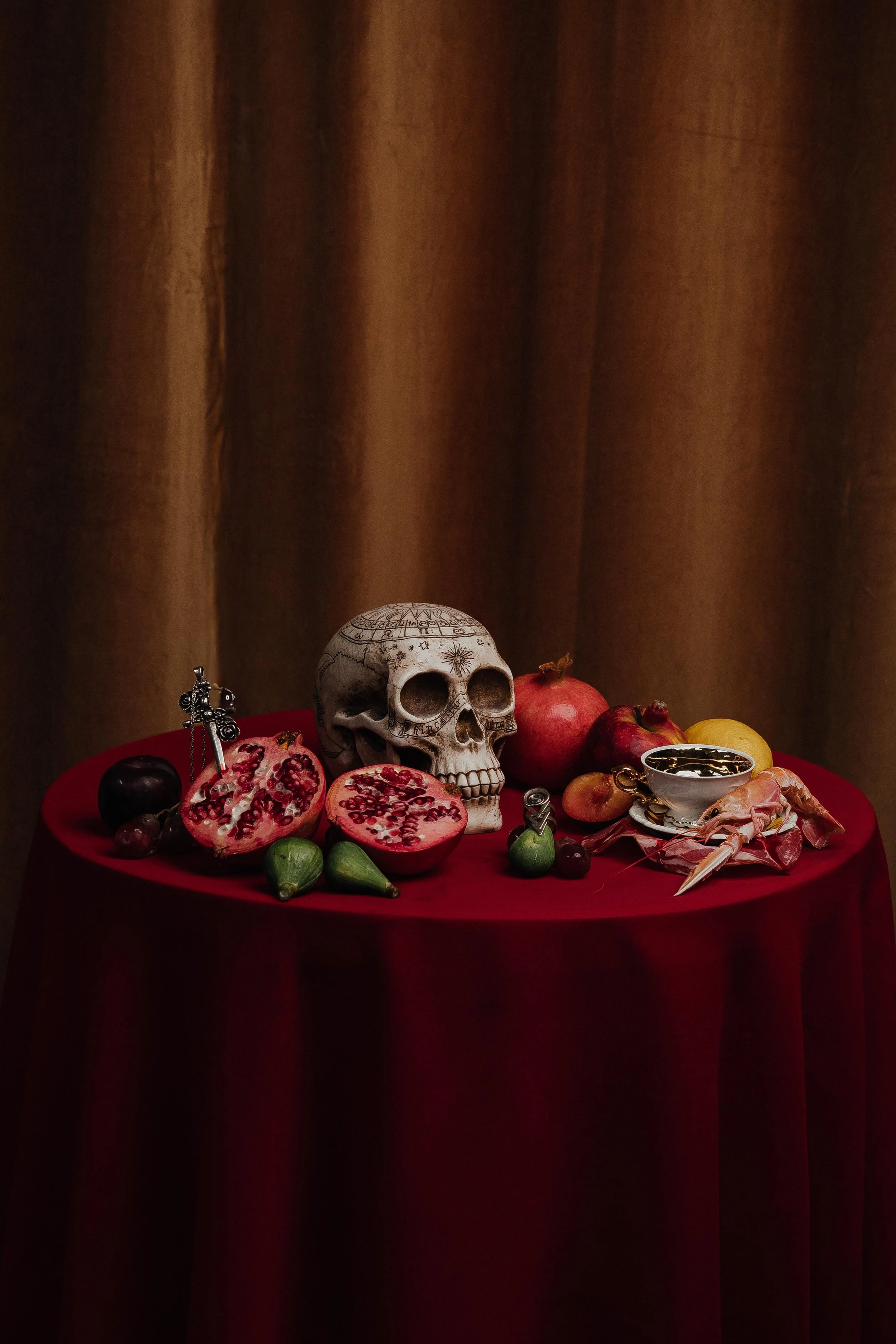 A still life arrangement of fruits, a skull, jewelry, and a goblet on a red tablecloth against a brown curtain background.