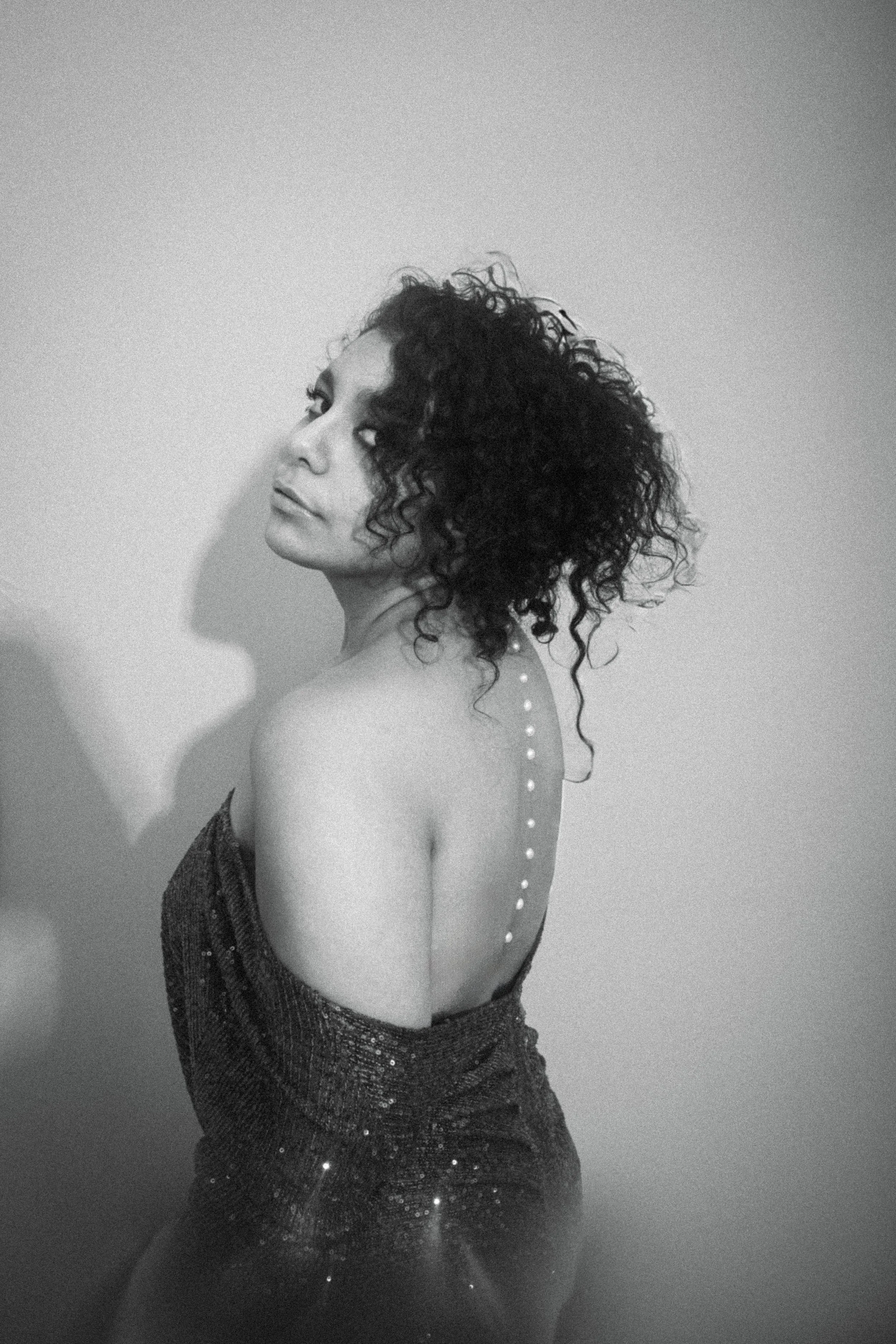 Black and white photo of a woman with curly hair, wearing a sparkly dress, turned slightly to her right, looking over her shoulder.