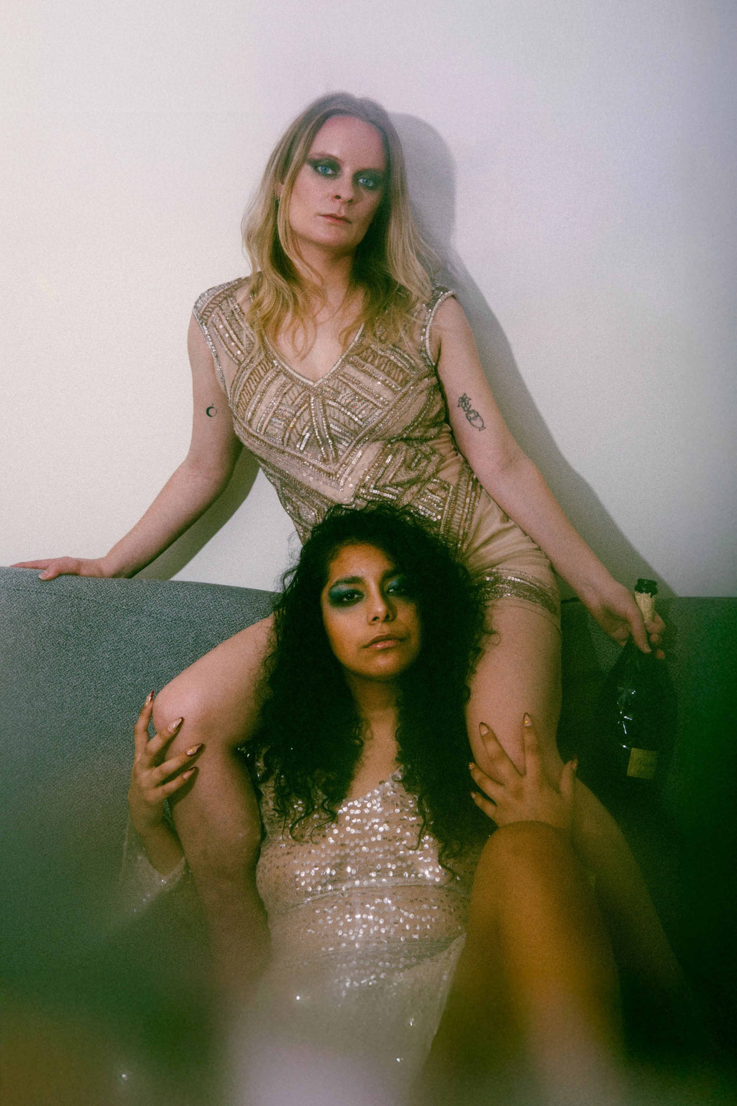 Two women with dark curly hair and bold eye makeup are in a playful pose. One woman is sitting on a sofa wearing a sequined dress, while the other woman is standing behind her, leaning on the wall, wearing a decorative dress with tattoos on her arms.