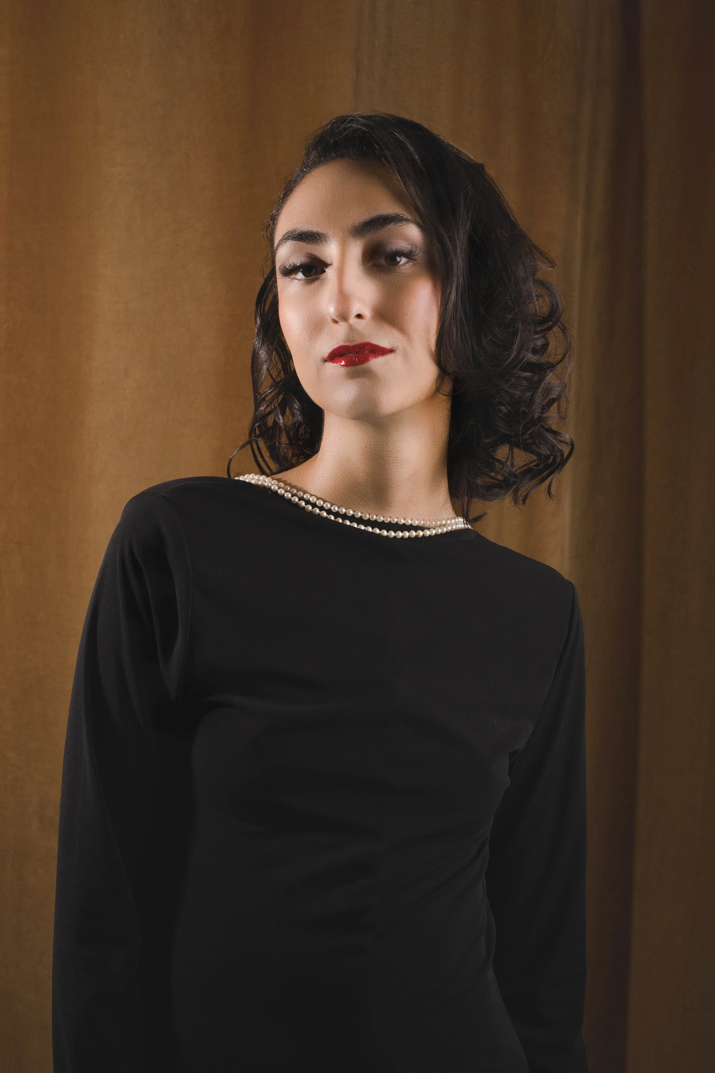 A woman with dark, curly hair wearing a black dress and a pearl necklace, posed in front of a brown curtain.