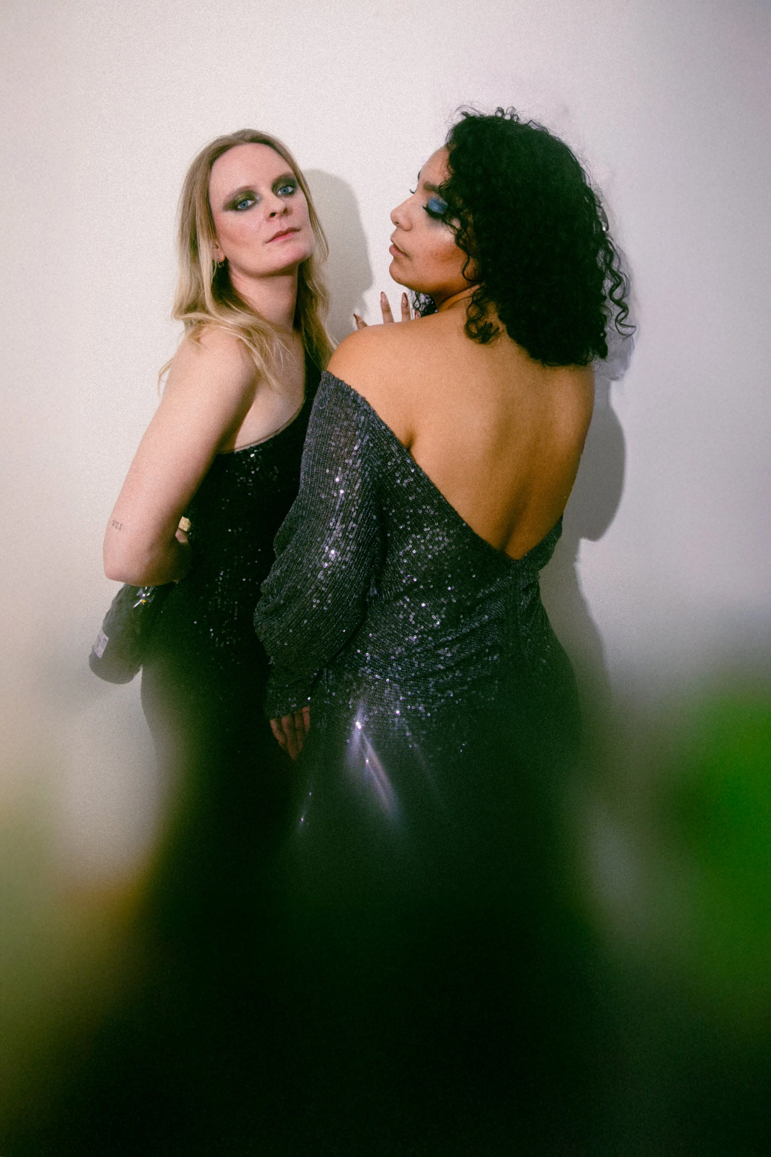 Two women with glamorous makeup and sparkly dresses standing against a plain white wall, one with blonde hair and the other with black curly hair.