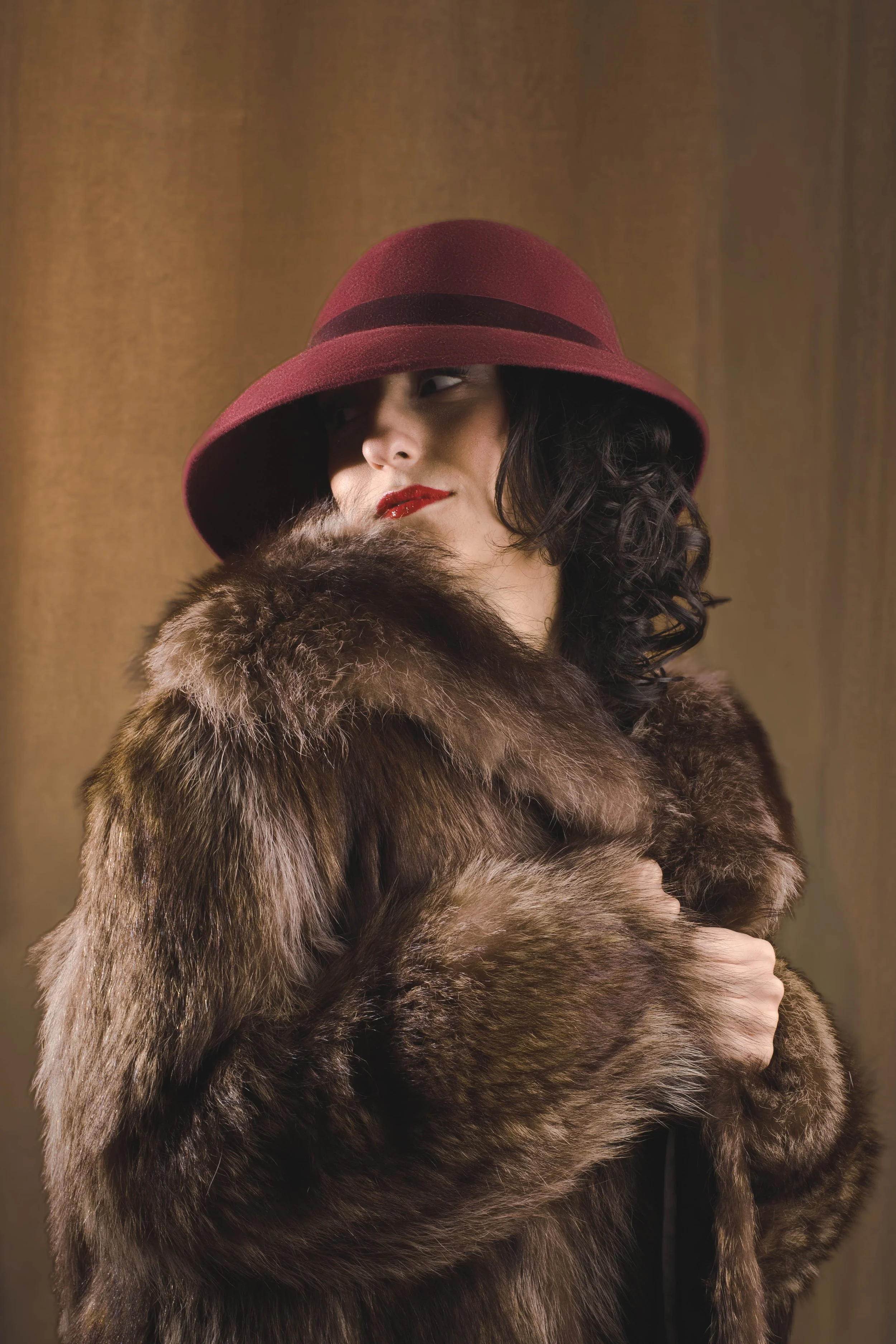A woman wearing a maroon hat with black band, red lipstick, and a fur coat, against a brown background.
