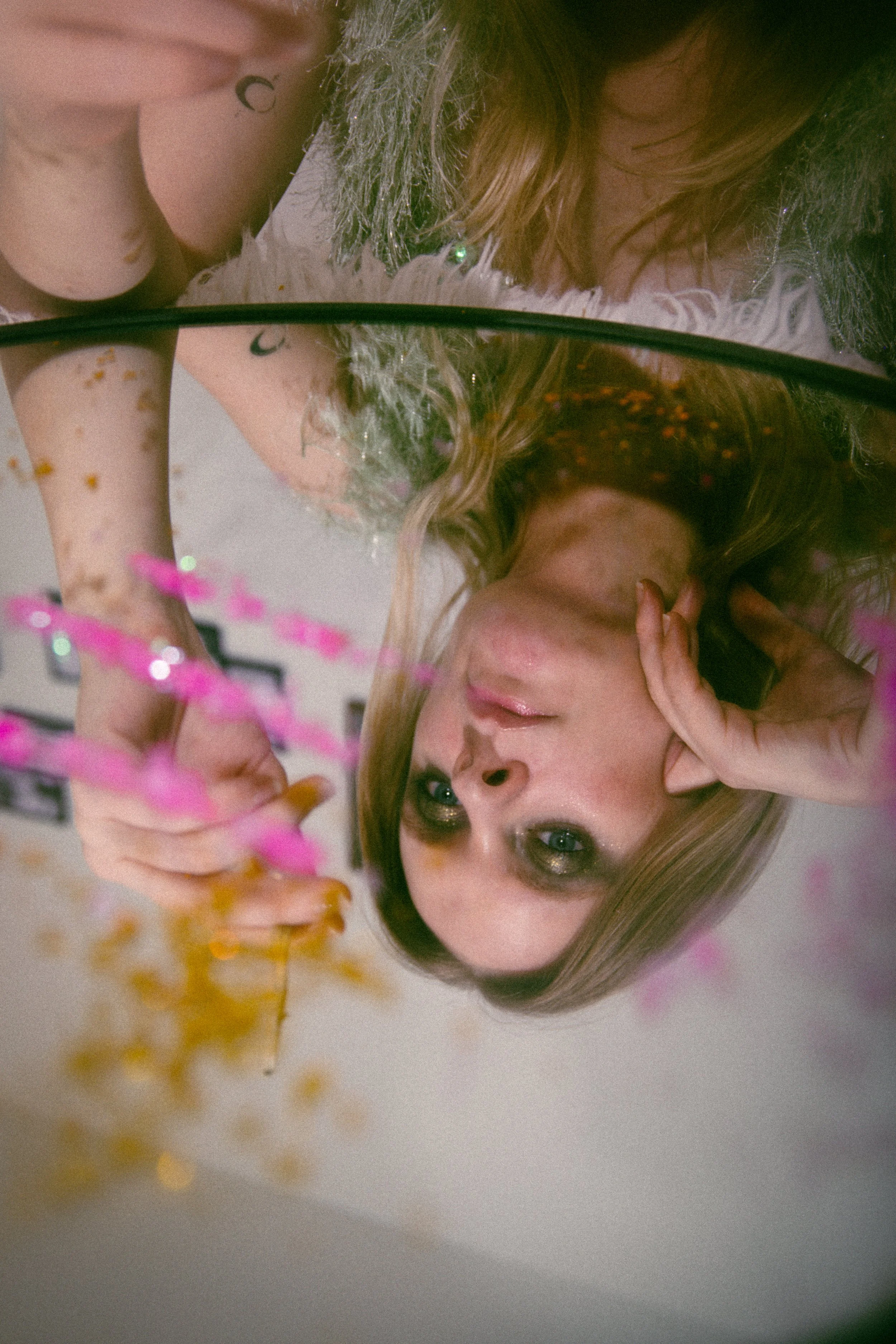 A young woman with green eyes and metallic eyeshadow takes a selfie in a round mirror, with her face resting on her hand, and pink and gold decorations are visible out of focus in the foreground.