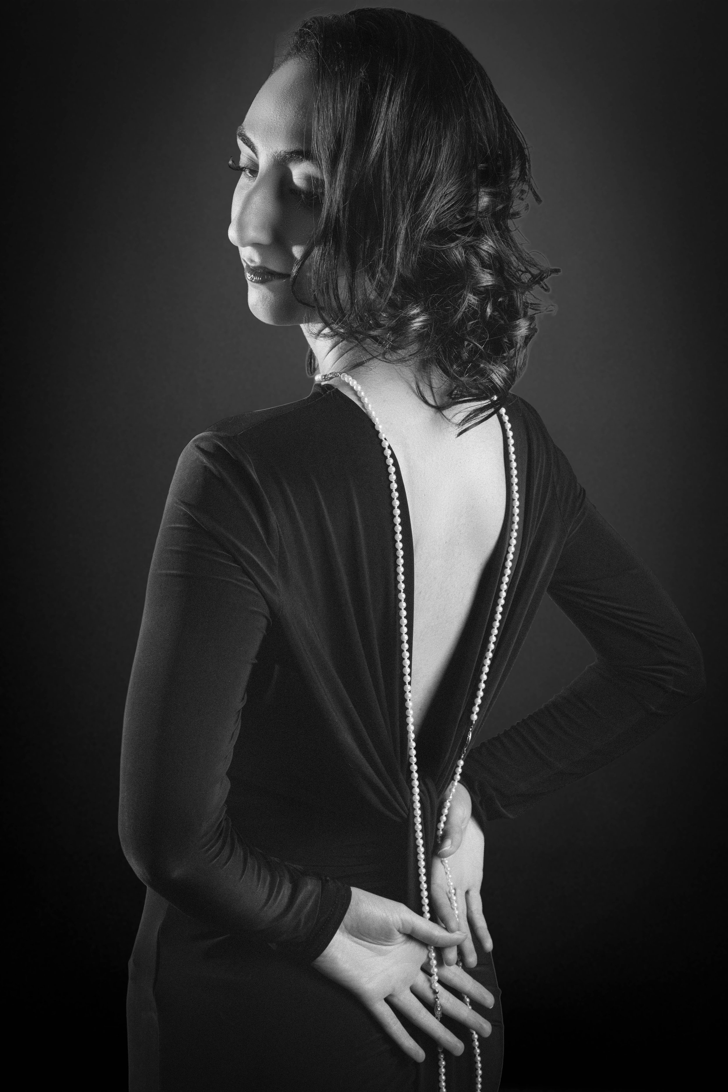 Black and white portrait of a woman with shoulder-length wavy hair, wearing dark clothing with a feathered collar and a long pearl necklace, posed with her eyes closed and hands on her waist against a dark background.