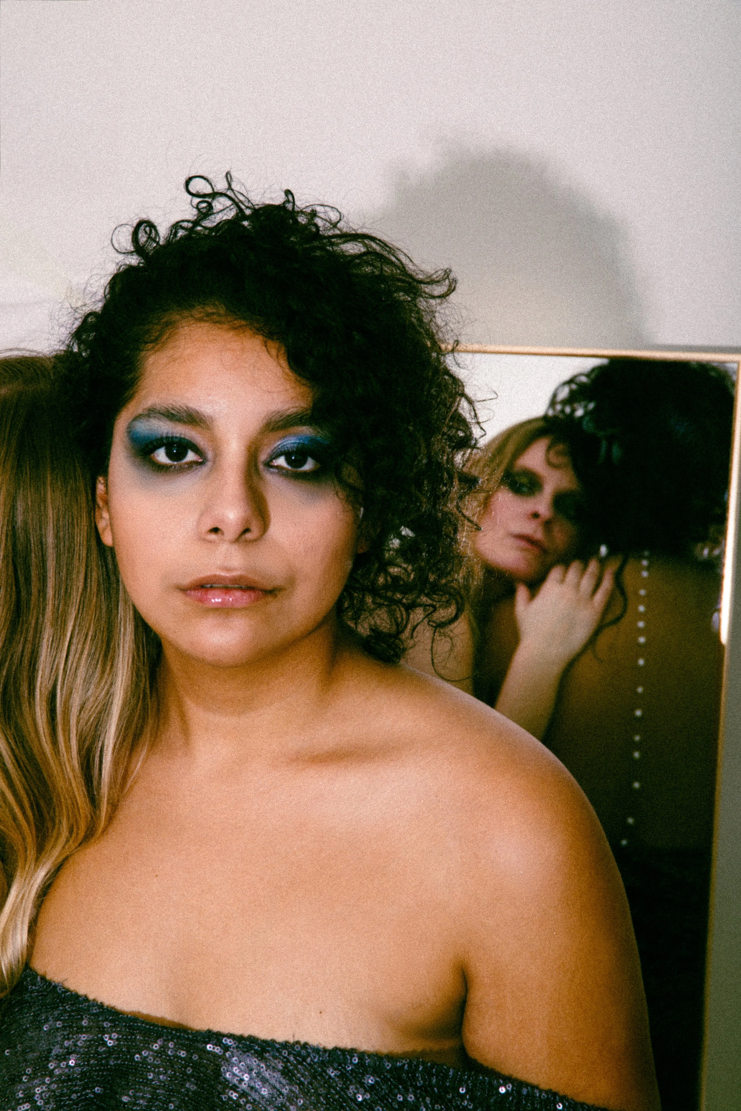 Close-up of a woman with curly dark hair and blue eyeshadow, wearing a black sequined top, standing in front of a mirror reflecting her face and part of her shoulder.
