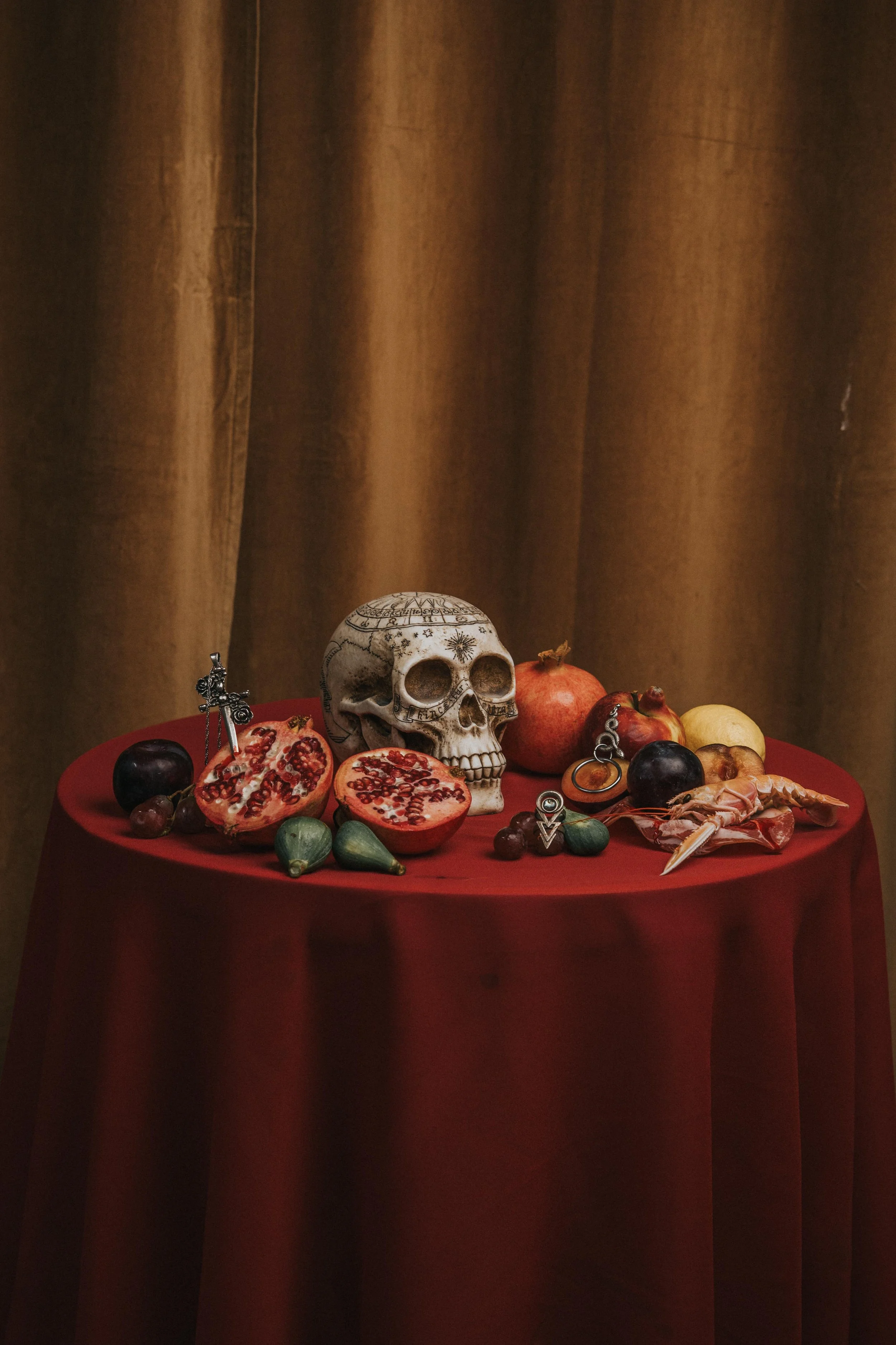A table with a skull, pomegranates, grapes, figs, apples, a small jewelry box, and silver jewelry, set against a brown curtain backdrop.