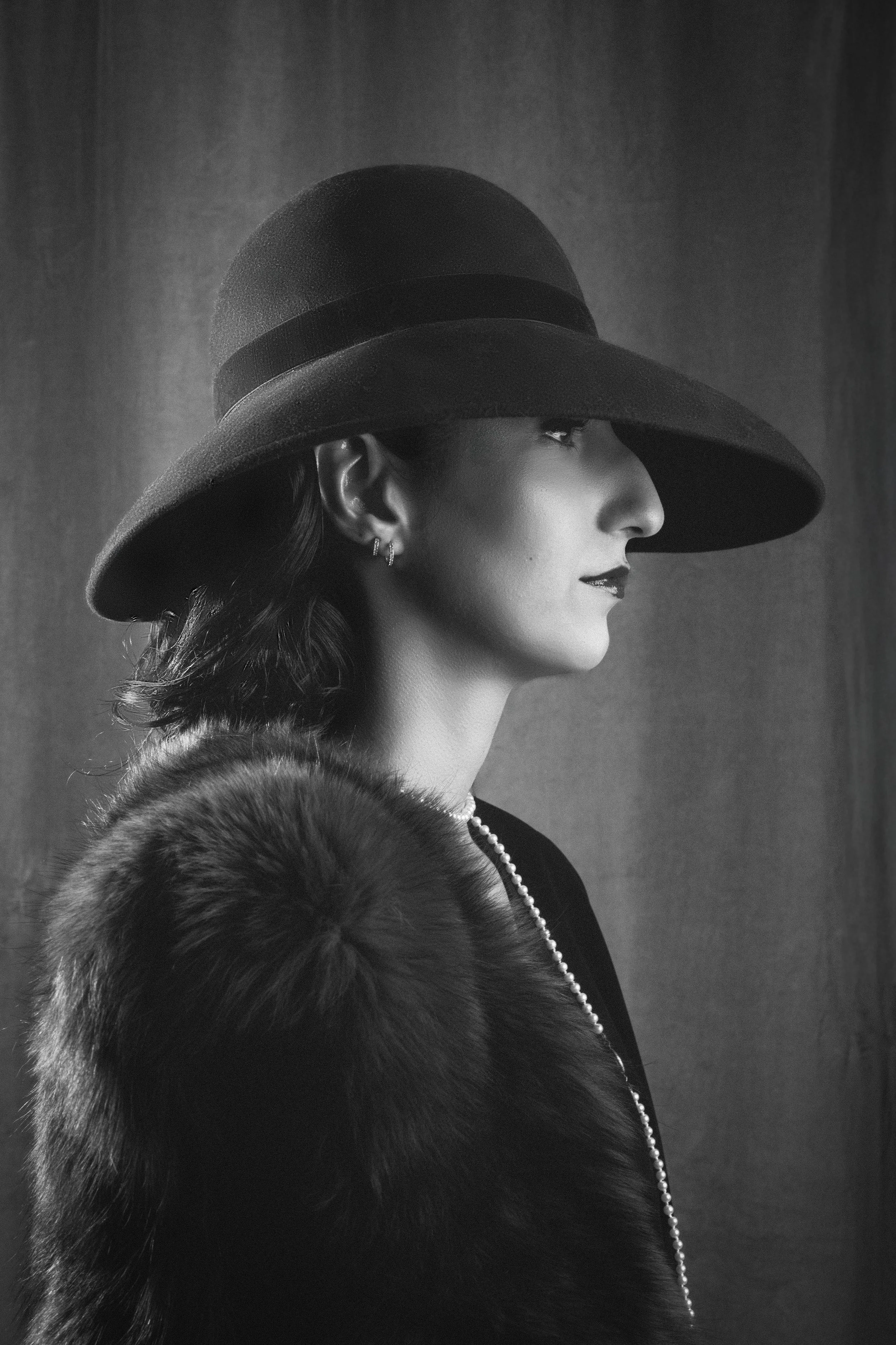 A black and white profile of a woman wearing a large, wide-brimmed hat, pearl necklace, earrings, and a fur stole.