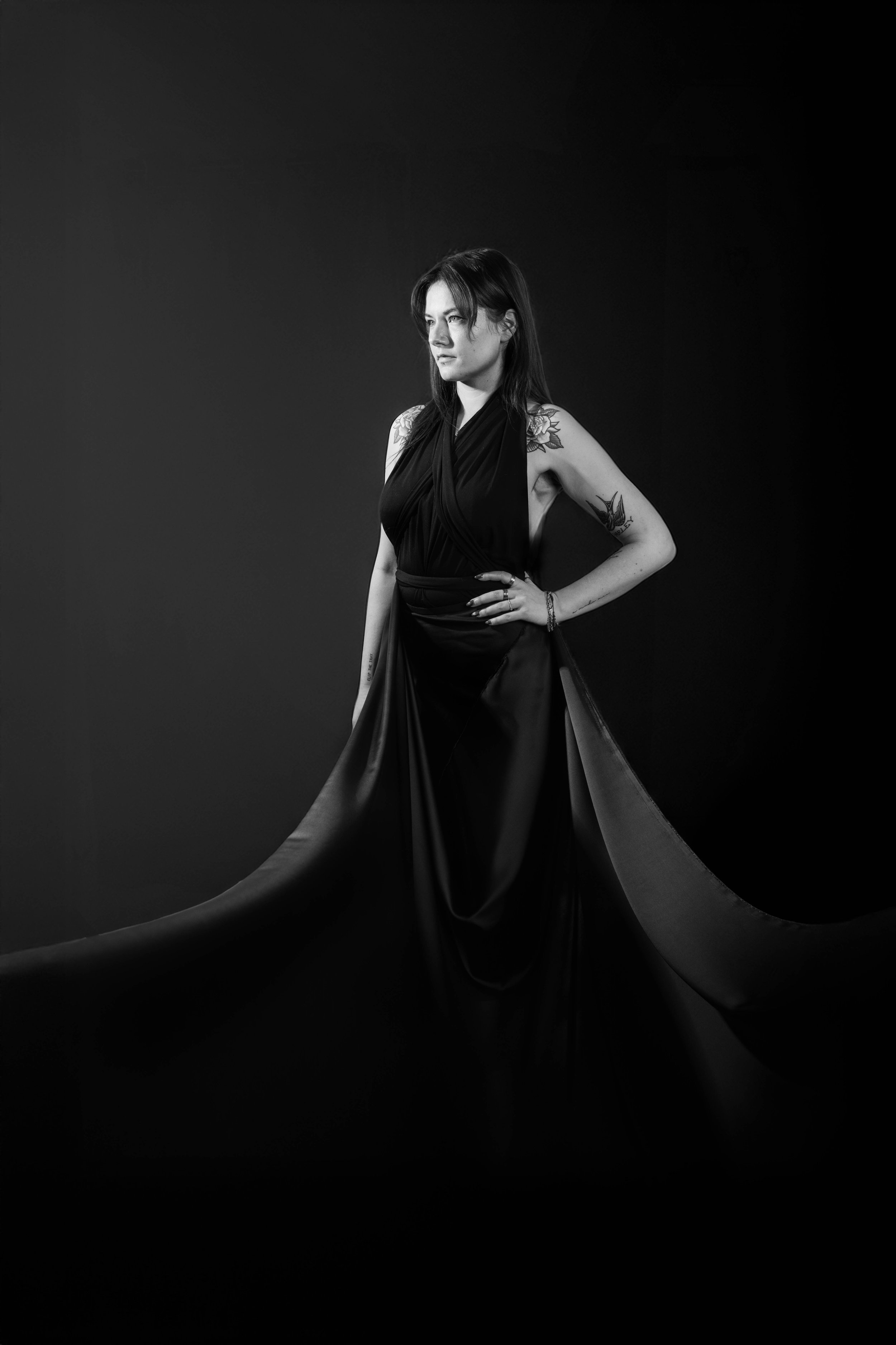 A black and white portrait of a woman with long dark hair, wearing a dark, flowing gown, standing against a dark background. She has tattoos on her arms and a contemplative expression.