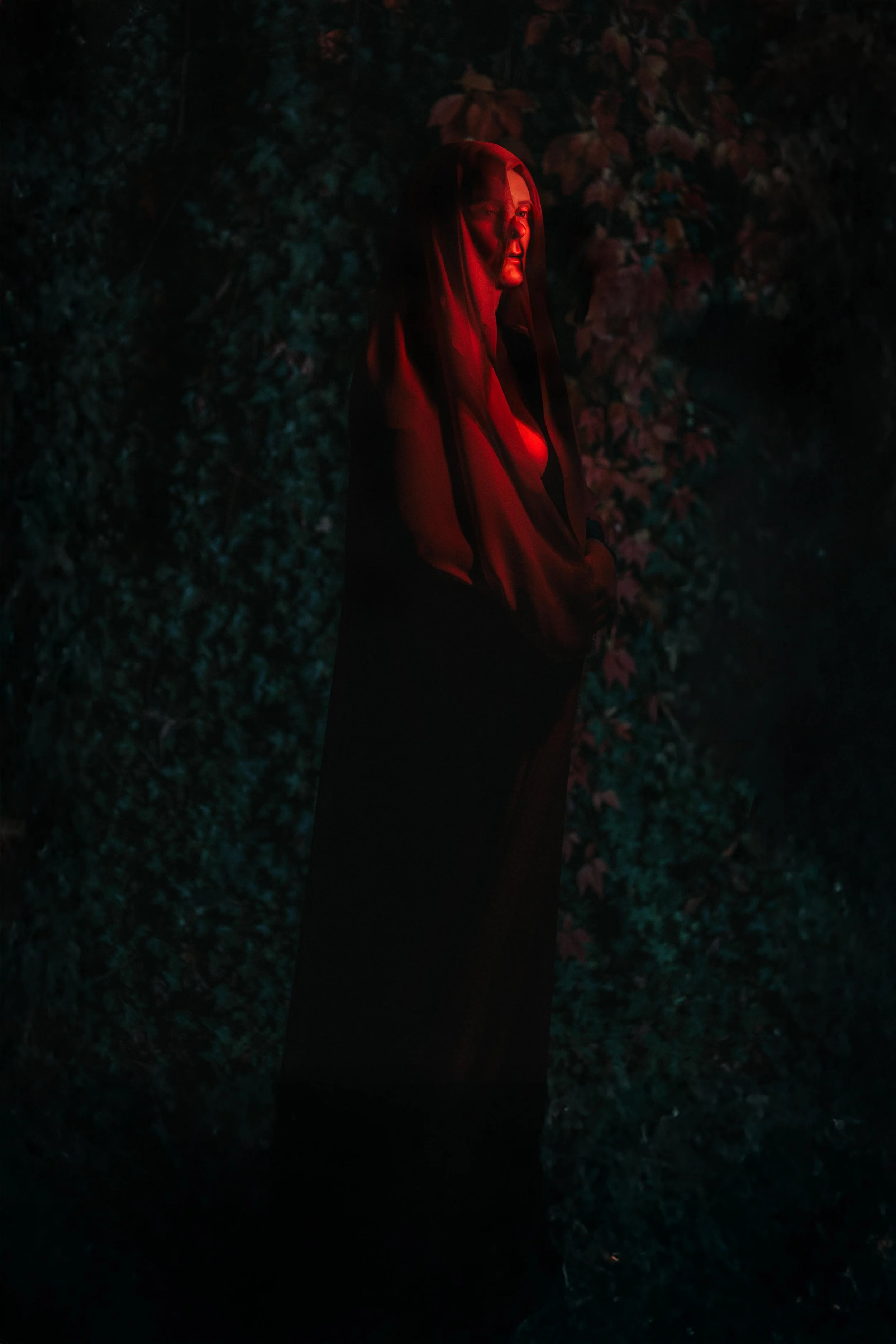 A woman standing outdoors near a wall of leaves, illuminated by red light, wearing a dark hoodie with a hood over her head.