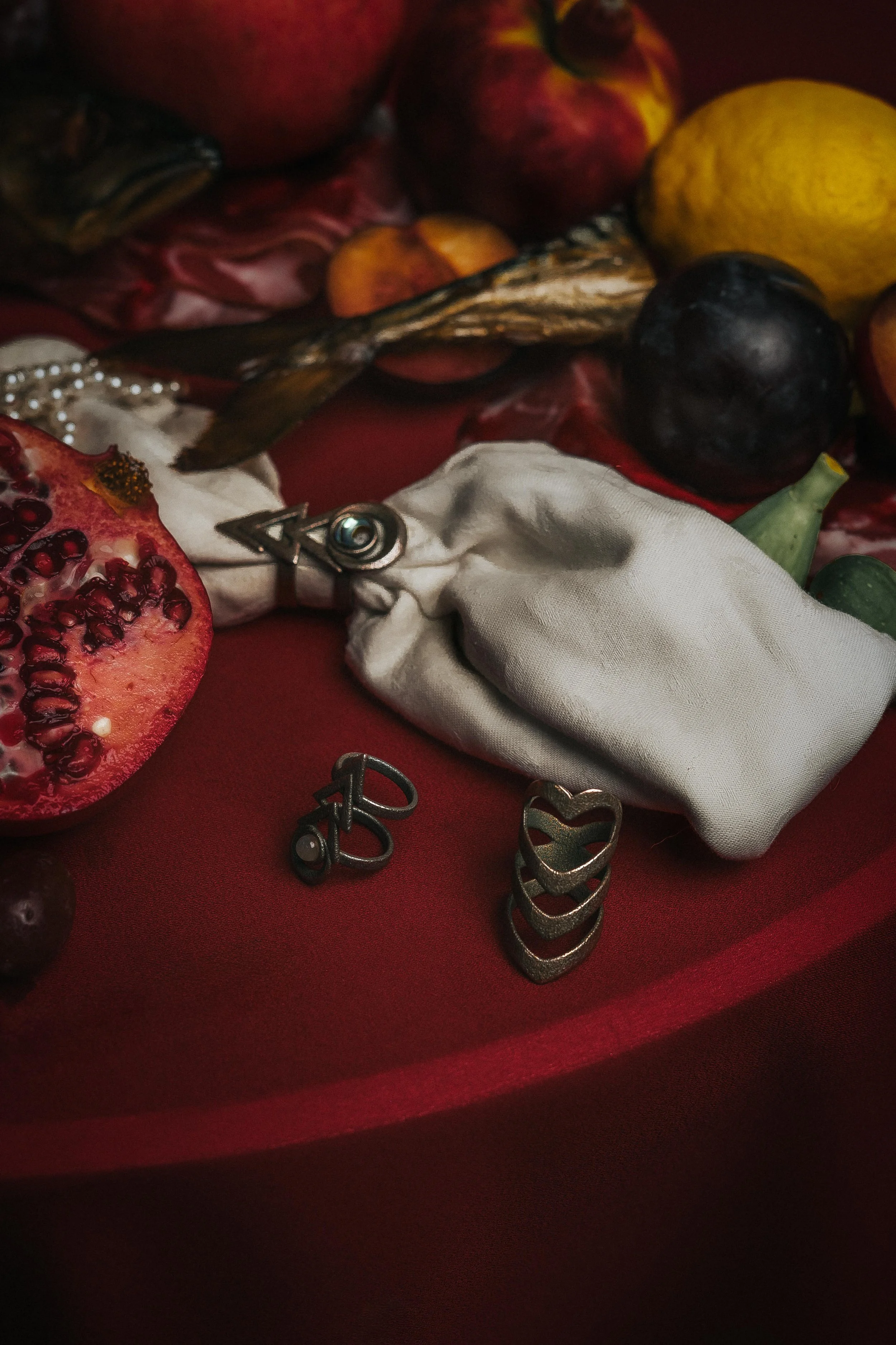 A collection of jewelry, including stacked heart-shaped rings, a pearl necklace, earrings, and a silver pin, arranged on a red fabric surface with fruits in the background.