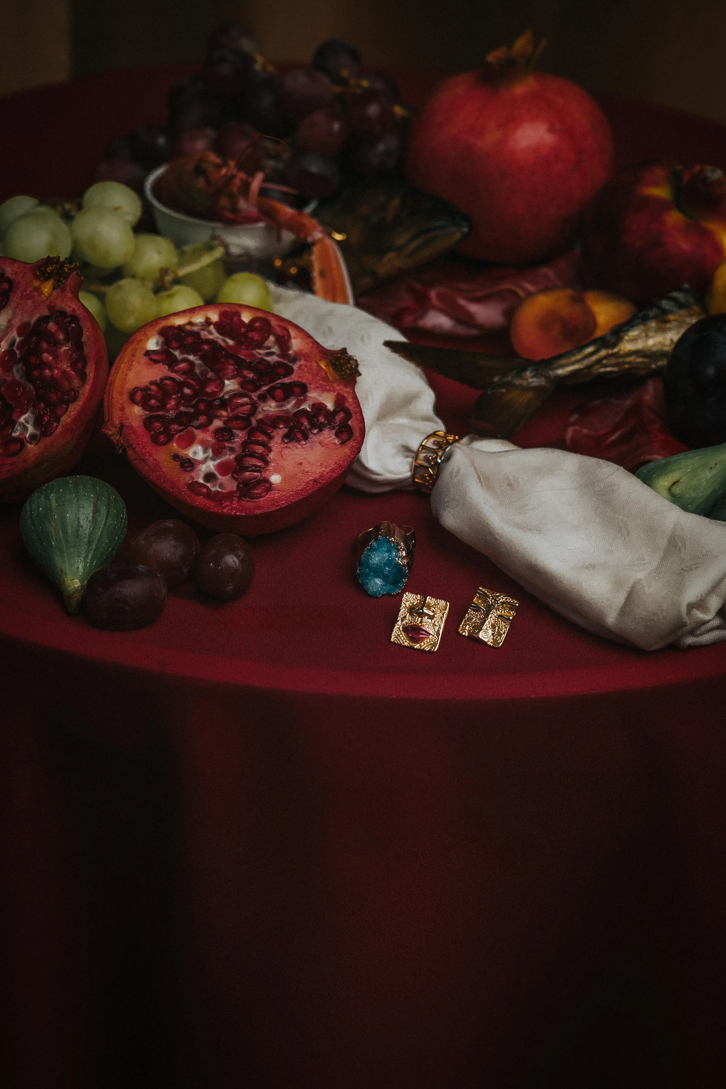 Close-up of a pomegranate cut in half, showing its red seeds, surrounded by grapes, apples, a fish, and jewelry on a red cloth.