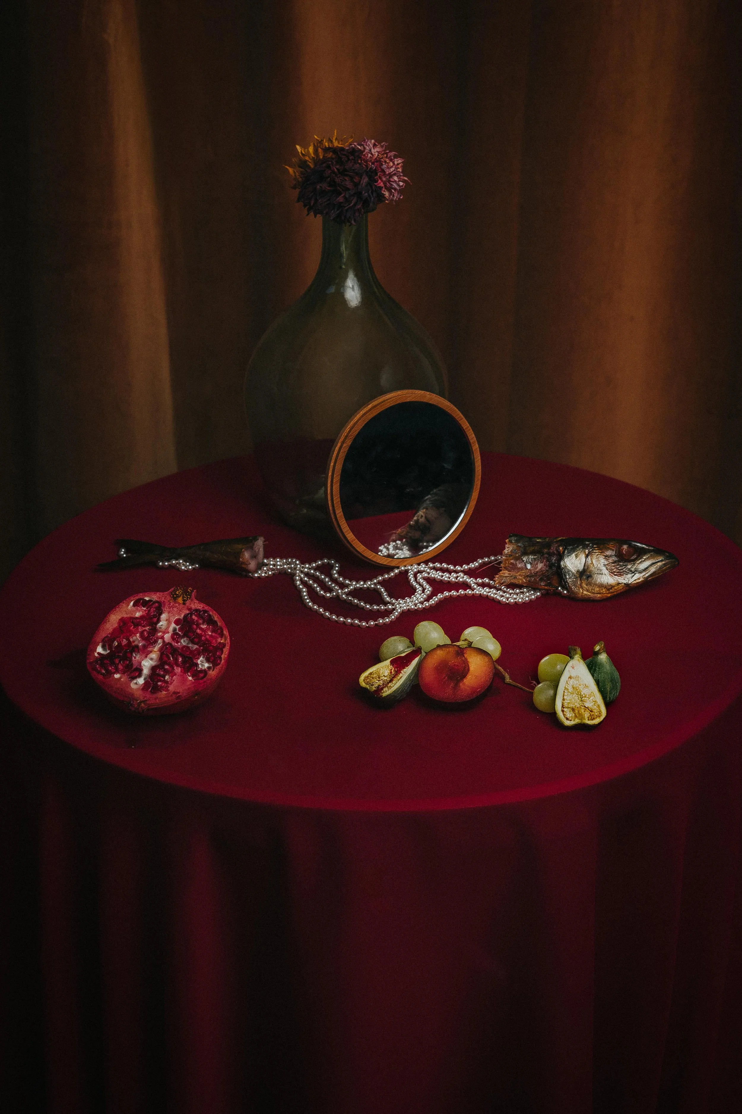 A dark red table with a large glass vase containing a purple dried flower, a clownfish, a mirror, grapes, an apple, a pear, a fig, and pomegranate halves. Renaissance inspired, painting