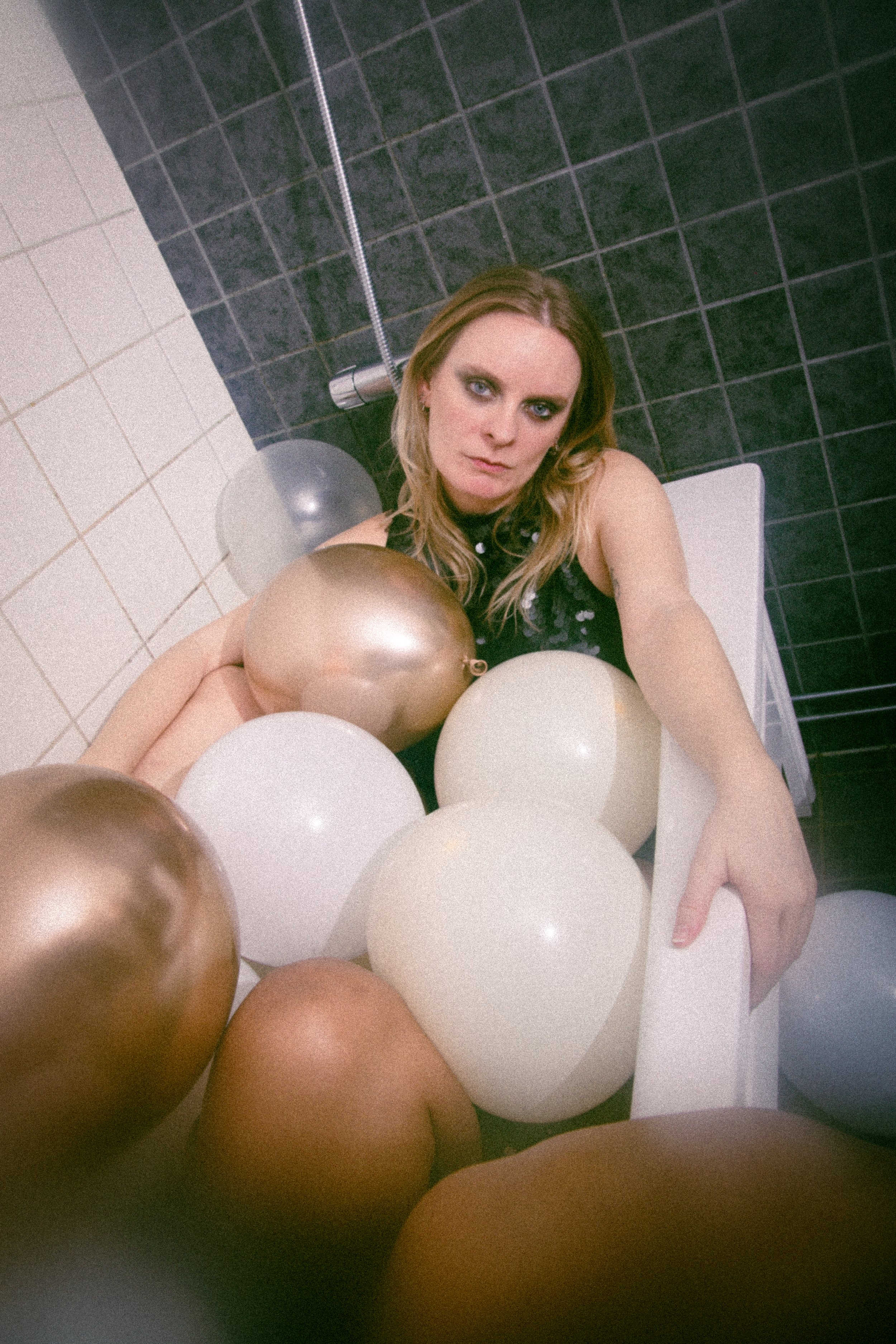 A woman with long blonde hair, blue eyes, and makeup sitting in a bathtub surrounded by metallic and white balloons in a bathroom with black and white tiled walls.
