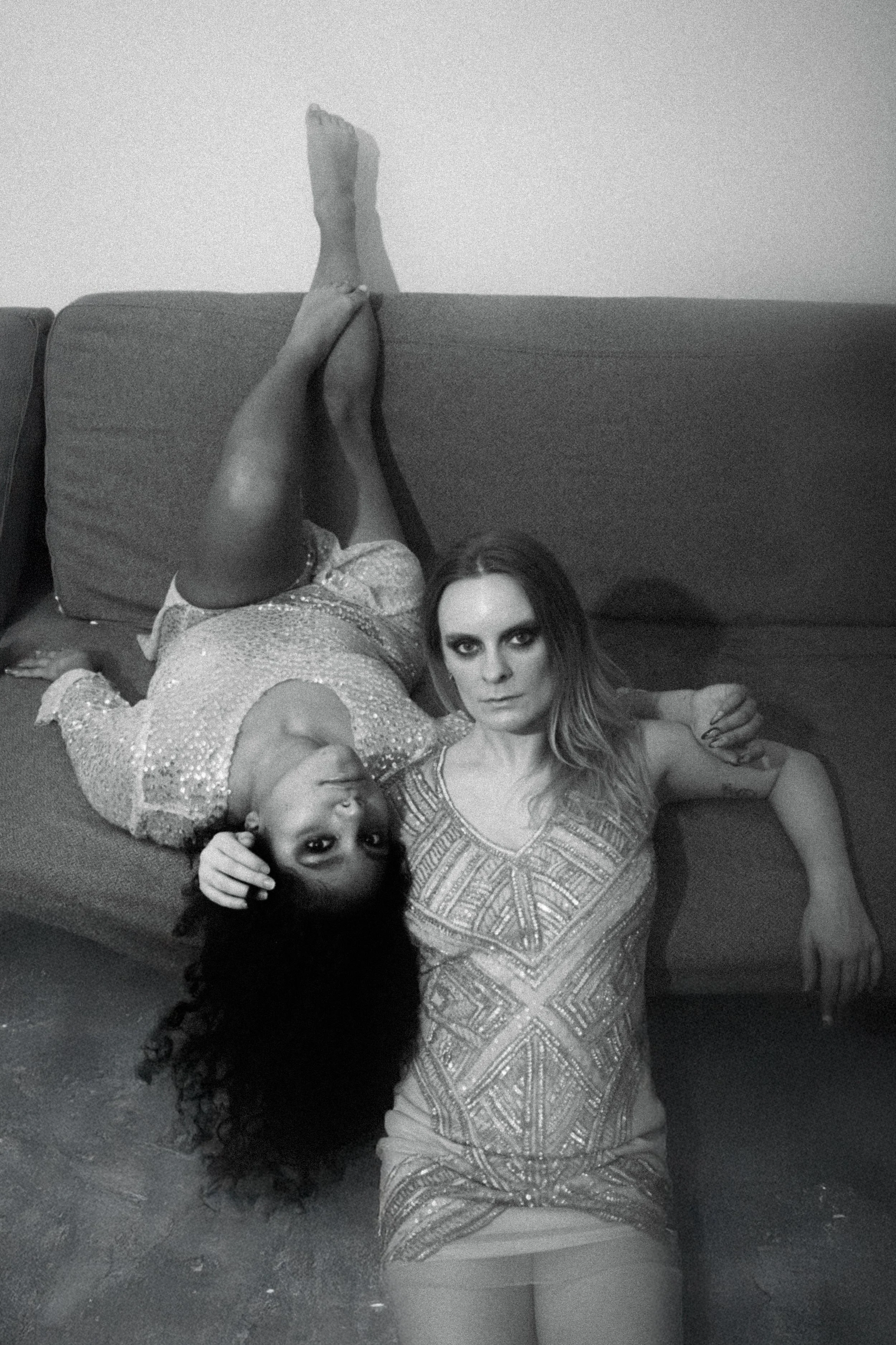 Black and white photo of two women resting on a sofa. One woman is lying on her side with her head resting on the other woman's arm, and the other woman is lying on her back with her head on the floor, looking at the camera. The woman lying on her si