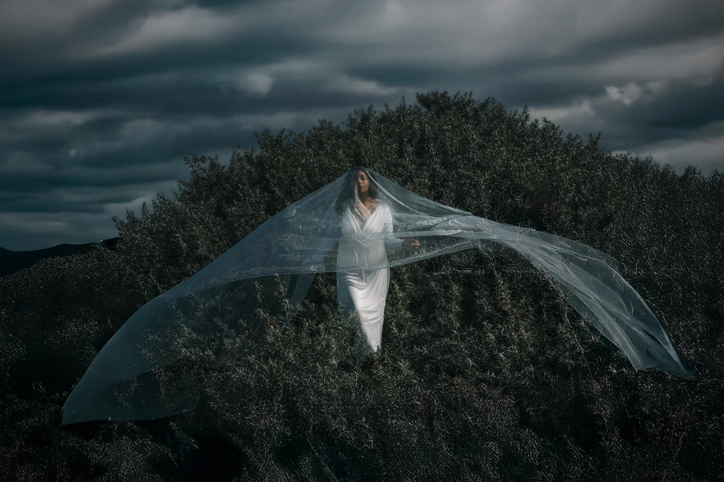 A woman dressed in white standing outdoors under a dark, cloudy sky, surrounded by trees, with a transparent fabric billowing around her.