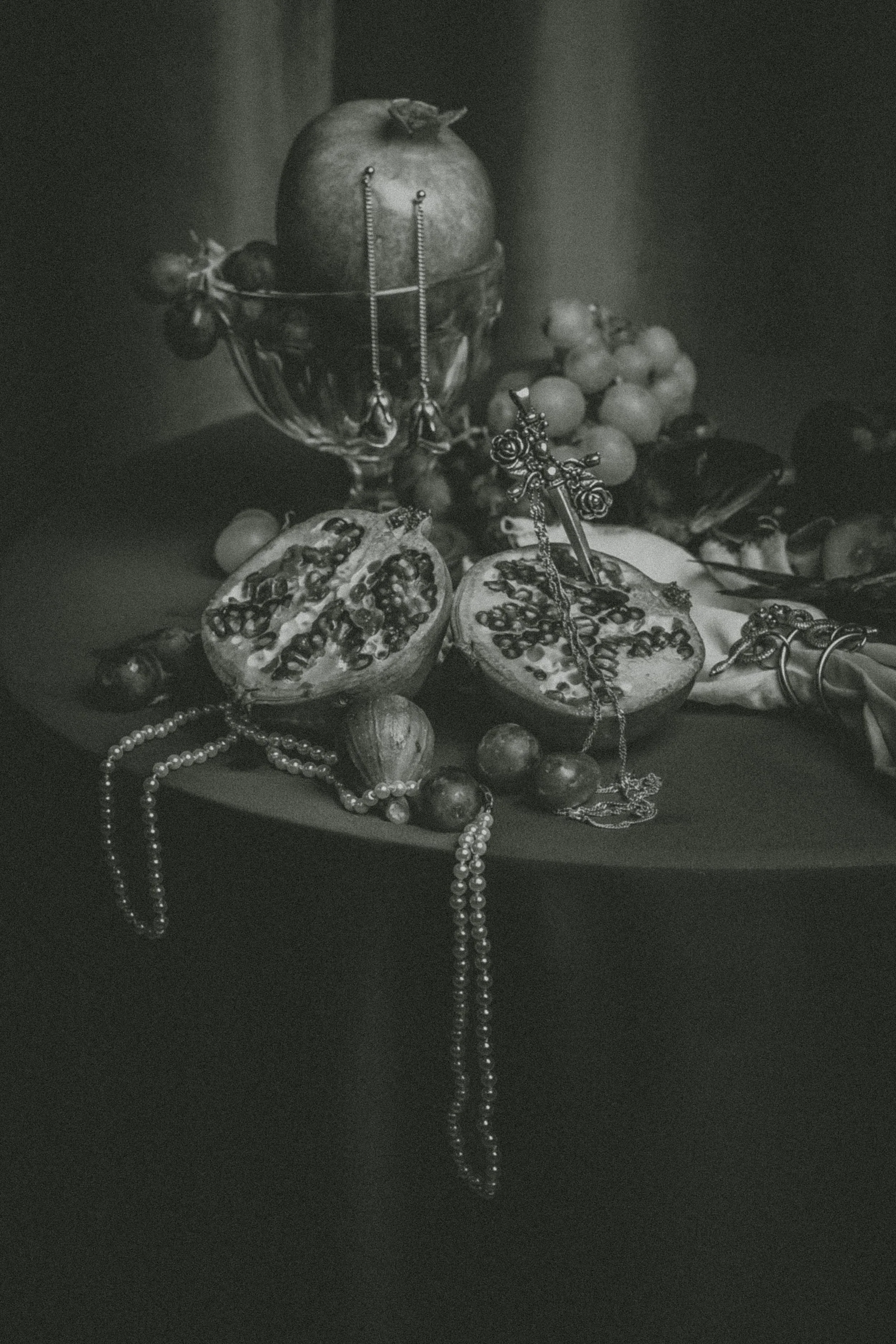 Black and white photograph of a still life arrangement with a pomegranate, grapes, jewelry, and cherries displayed in a glass bowl and scattered around, with some items hanging over the edge.