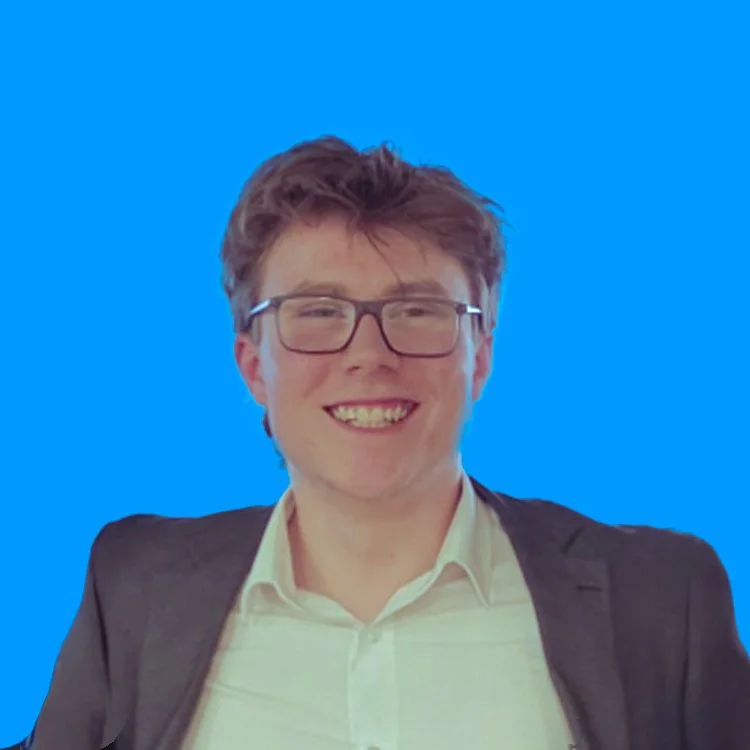 A young man with glasses and short brown hair, smiling and wearing a white shirt and dark blazer against a bright blue background.
