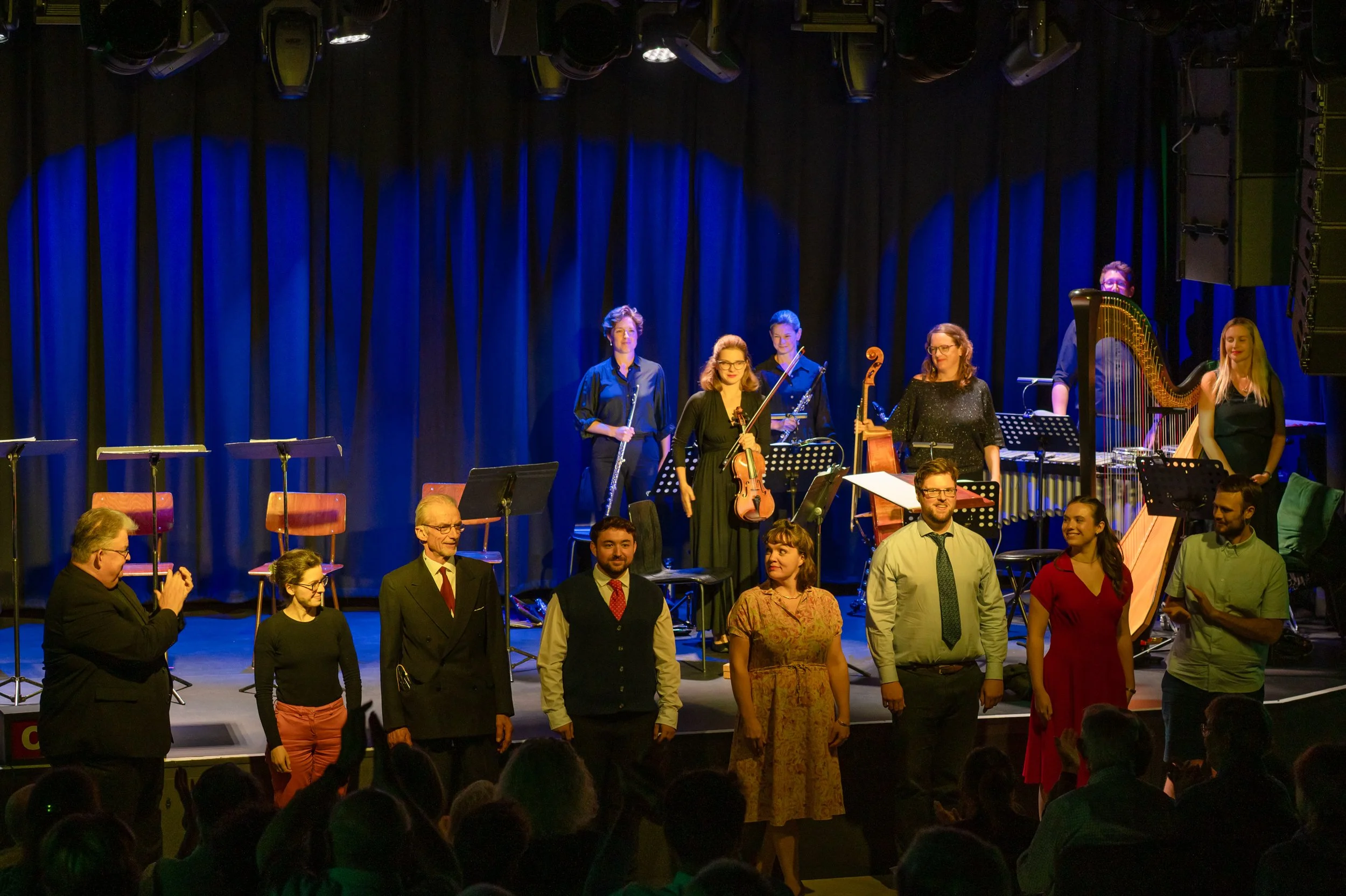 Orchestra members standing on stage after a performance with blue stage curtains and audience in front.