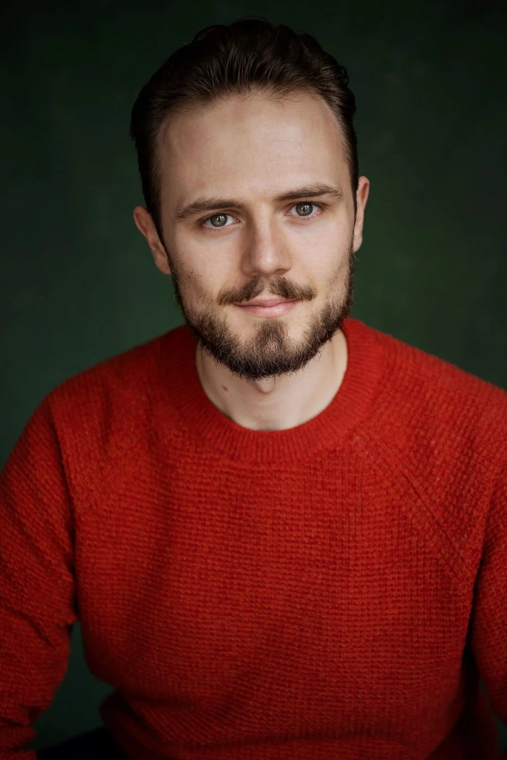 A young man with a beard and light skin, wearing a red sweater, smiling softly, against a dark green background.