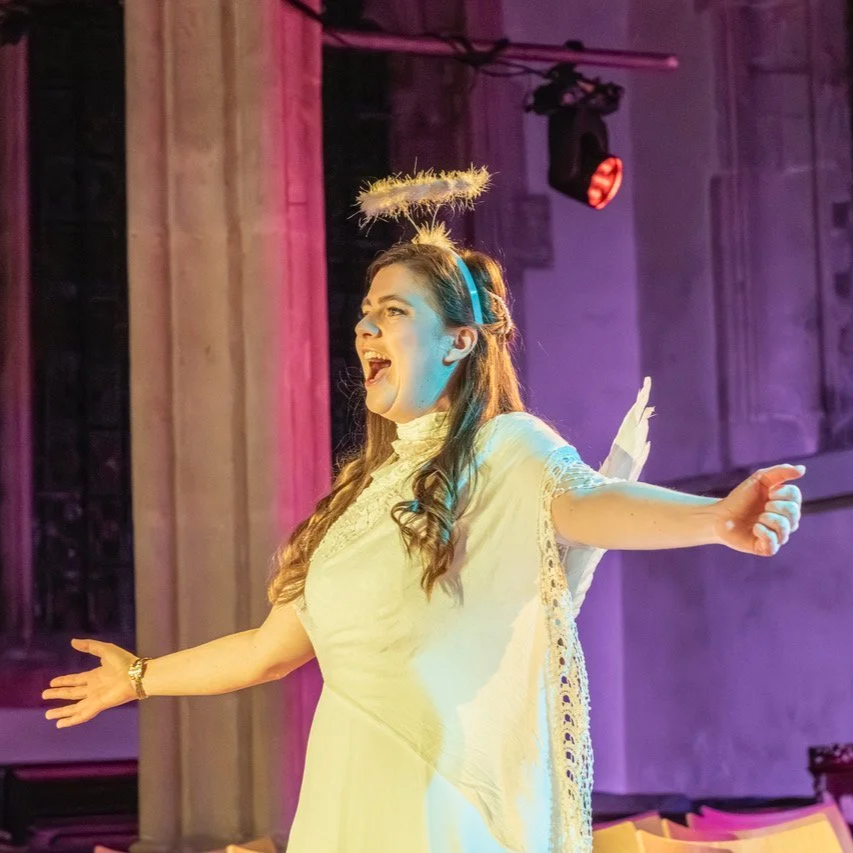 A woman dressed as an angel, with wings and a halo, performing on stage with her arms outstretched and an expression of joy, illuminated by stage lighting.