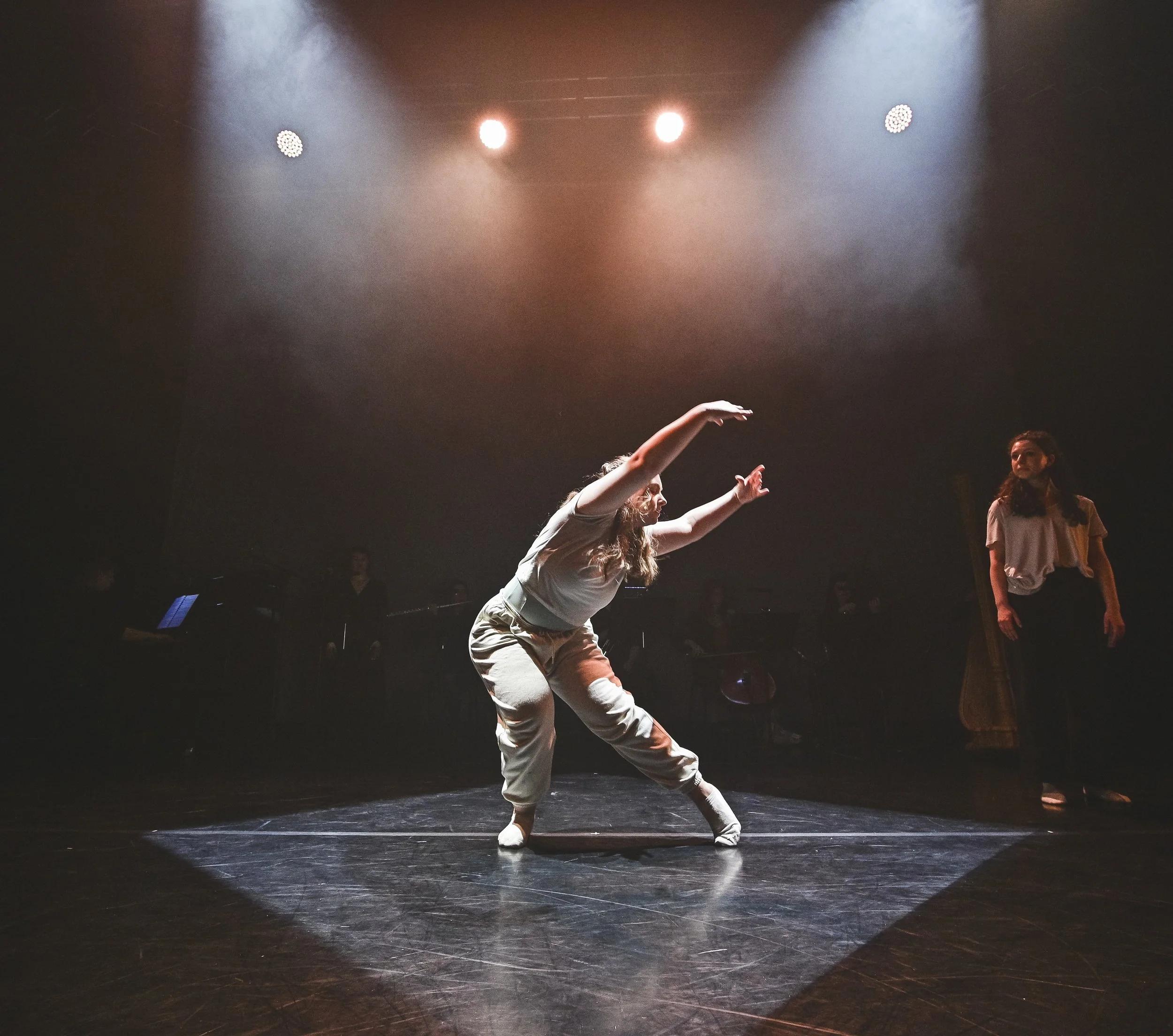 A dancer performs on stage under spotlight, wearing casual clothing and balancing on one leg with arms extended, with two women in the background watching.