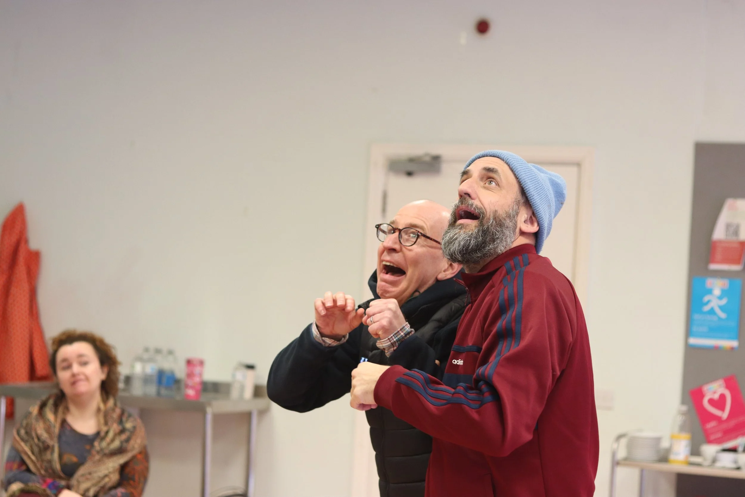 Two men, one wearing glasses and the other wearing a blue beanie, are standing indoors with their mouths open, looking upward. A woman with curly hair and a patterned scarf is seated in the background, and the room appears to be some kind of community or medical center with informational posters on the walls.