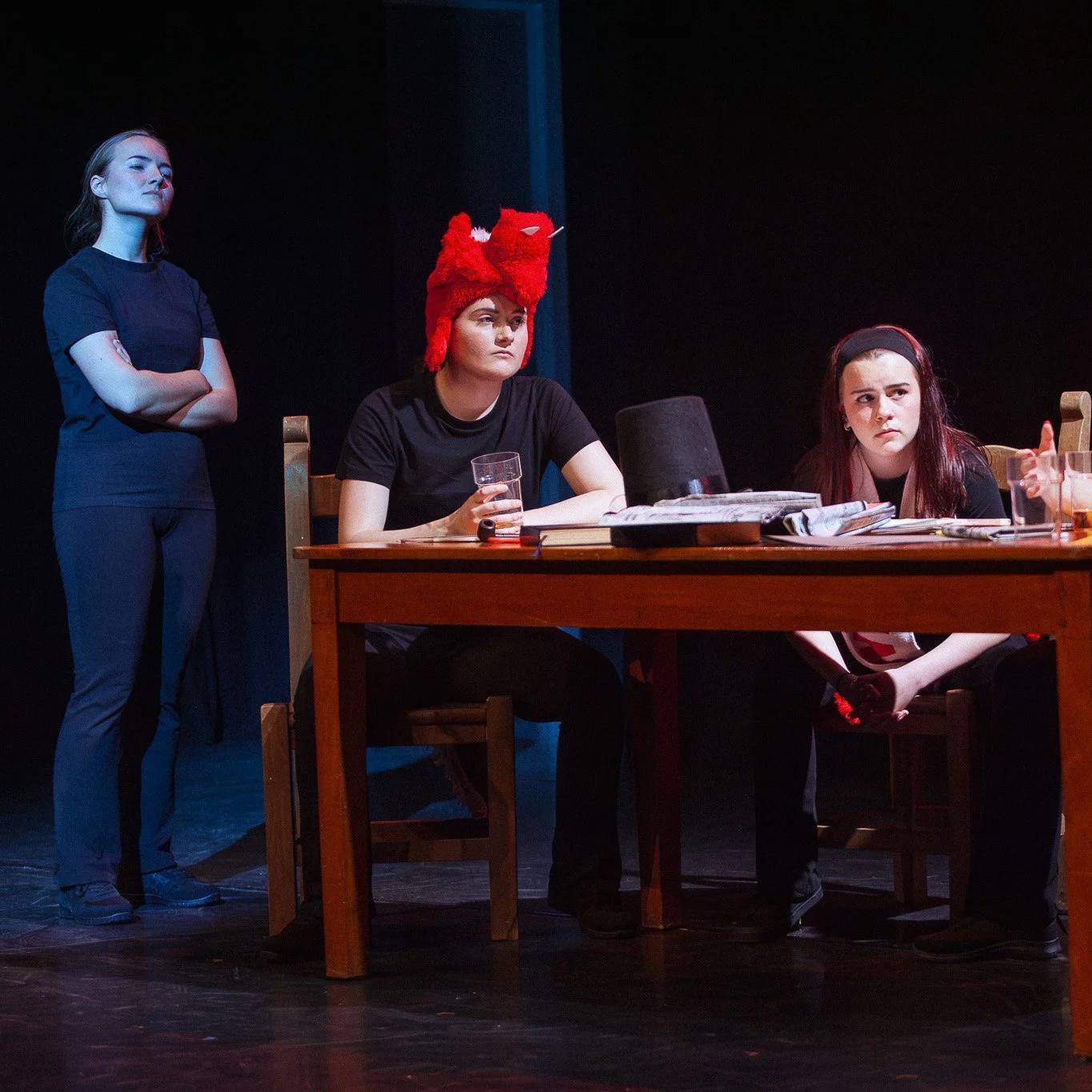 Three women in a theater scene. Two are seated at a table, one with a red fox hat, the other with a headband, both looking serious. The third woman stands with arms crossed, observing.