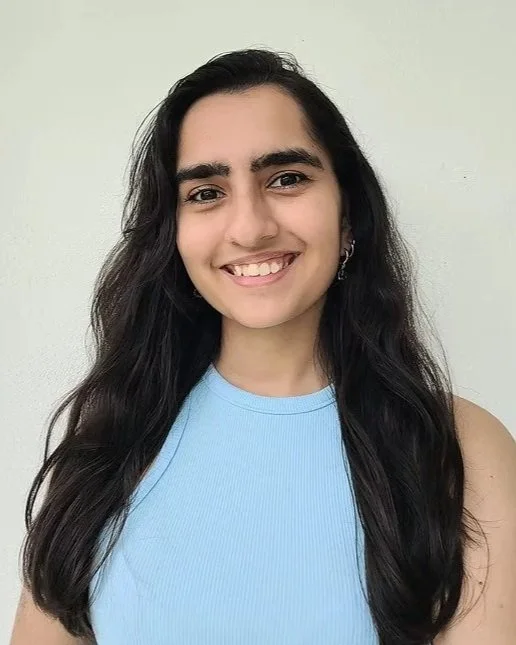 A young woman with long black hair, wearing a light blue sleeveless top, smiling, against a plain off-white wall.