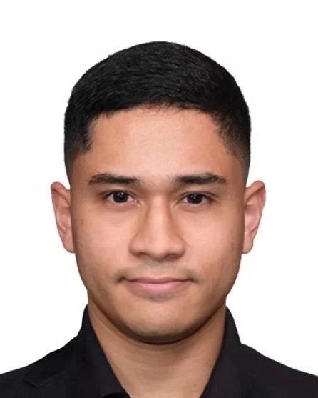 A young man with short black hair and a clean-shaven face, wearing a black shirt, looking directly at the camera against a plain white background.