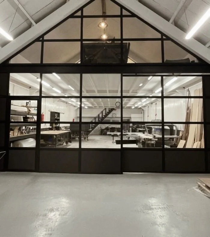 Binnen in een workshop of atelier met grote ramen, werkbanken, machines en hout. Sommige materialen staan op een plank en er is een trap naar een bovenverdieping.