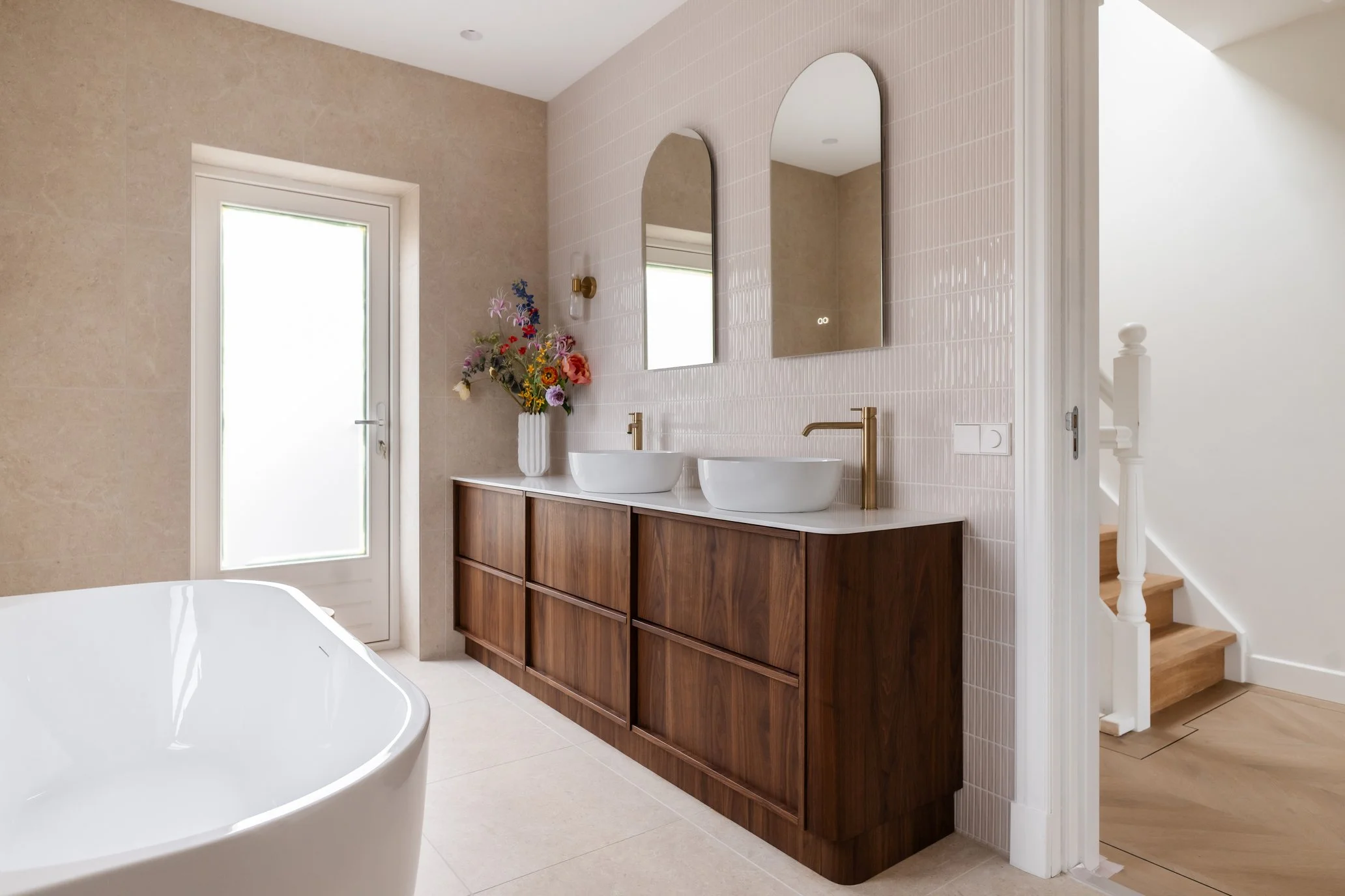 Moderne badkamer met dubbele wastafel, twee ronde witte wasbakken, gouden kranen, een houten kast onder de wastafels, tweemaal een spiegel aan de muur, een vaas met bloemen, een raam, een trap en een witte badkuip in de voorgrond.