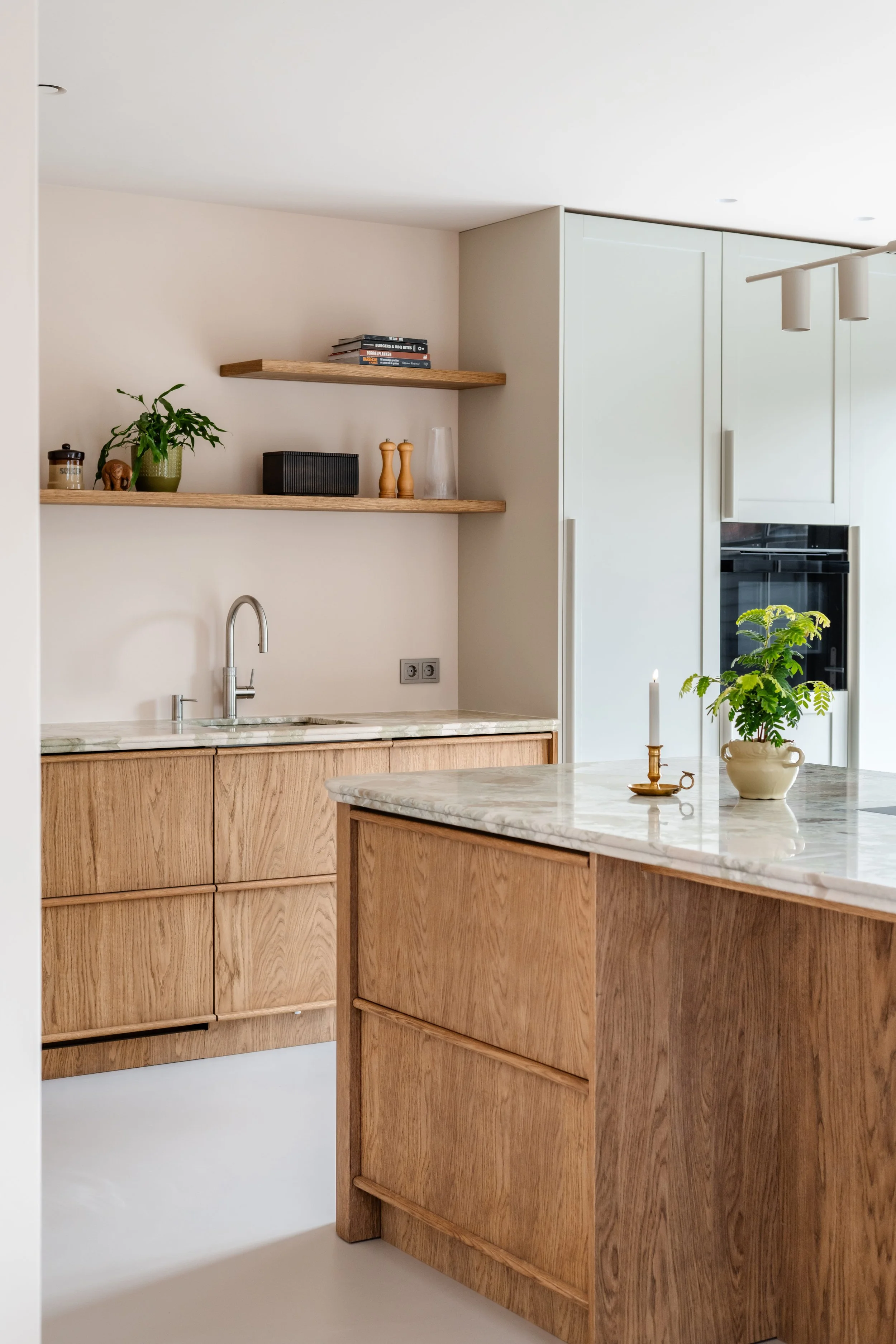 Moderne keuken met houten kasten en een wit marmeren aanrecht. Open planken met boeken en decoratieve items. Groene plant en kaarsen op het eiland.