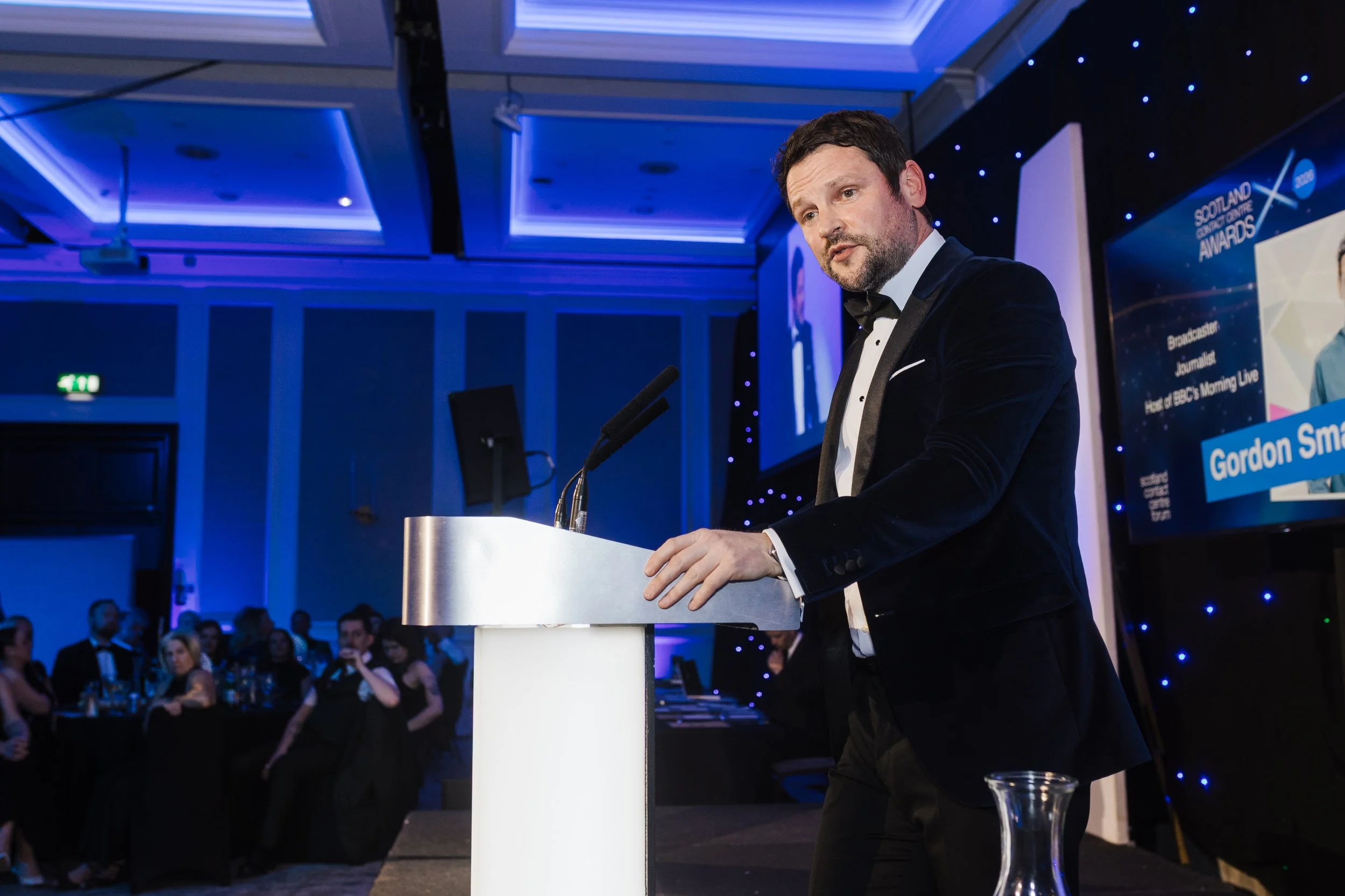 Man in tuxedo speaking at podium during awards ceremony with audience and large screen displaying event information in background.