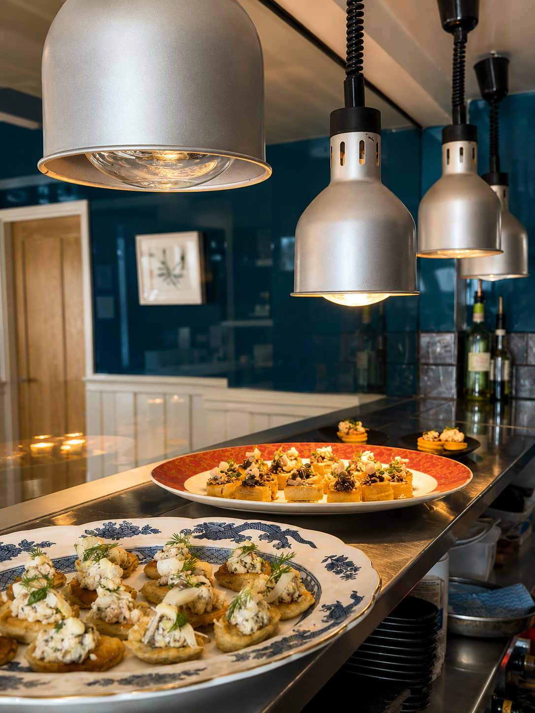 Close-up of appetizers on decorative plates under hanging industrial-style pendant lights in a restaurant kitchen.