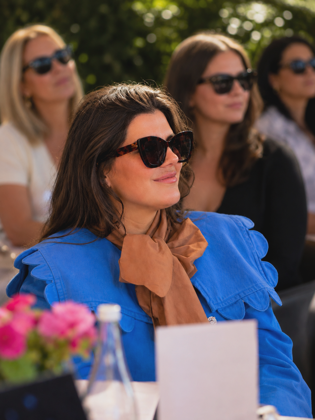 A woman with long brown hair wearing large tortoiseshell sunglasses, a bright blue jacket, and a brown scarf tied around her neck, sitting outdoors at a table with pink flowers and a glass bottle, attending an event with other women in the background also wearing sunglasses.