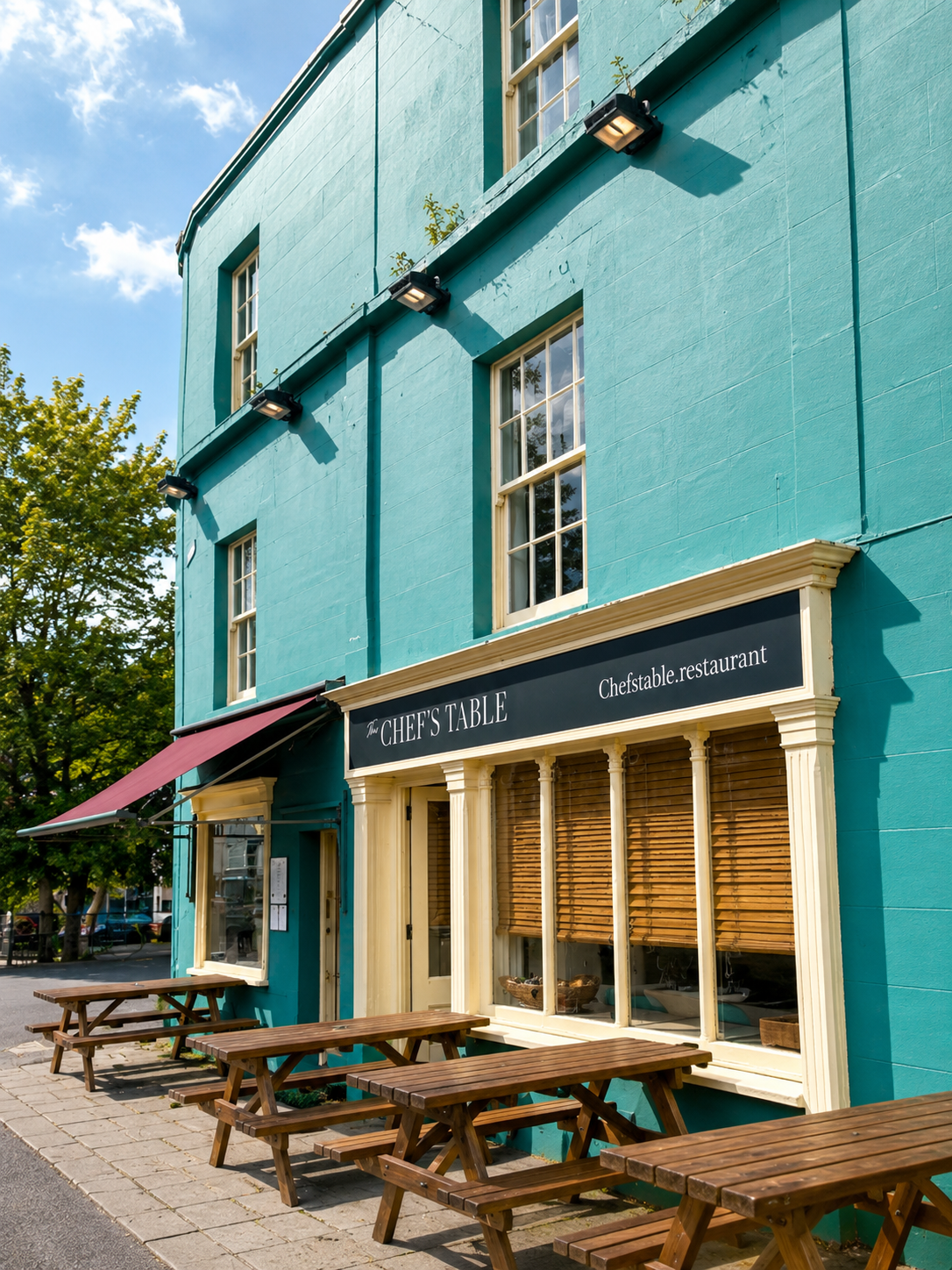 The exterior of a teal-colored restaurant building with outdoor wooden picnic tables and benches, a black awning, and a sign reading 'The Chef's Table'.
