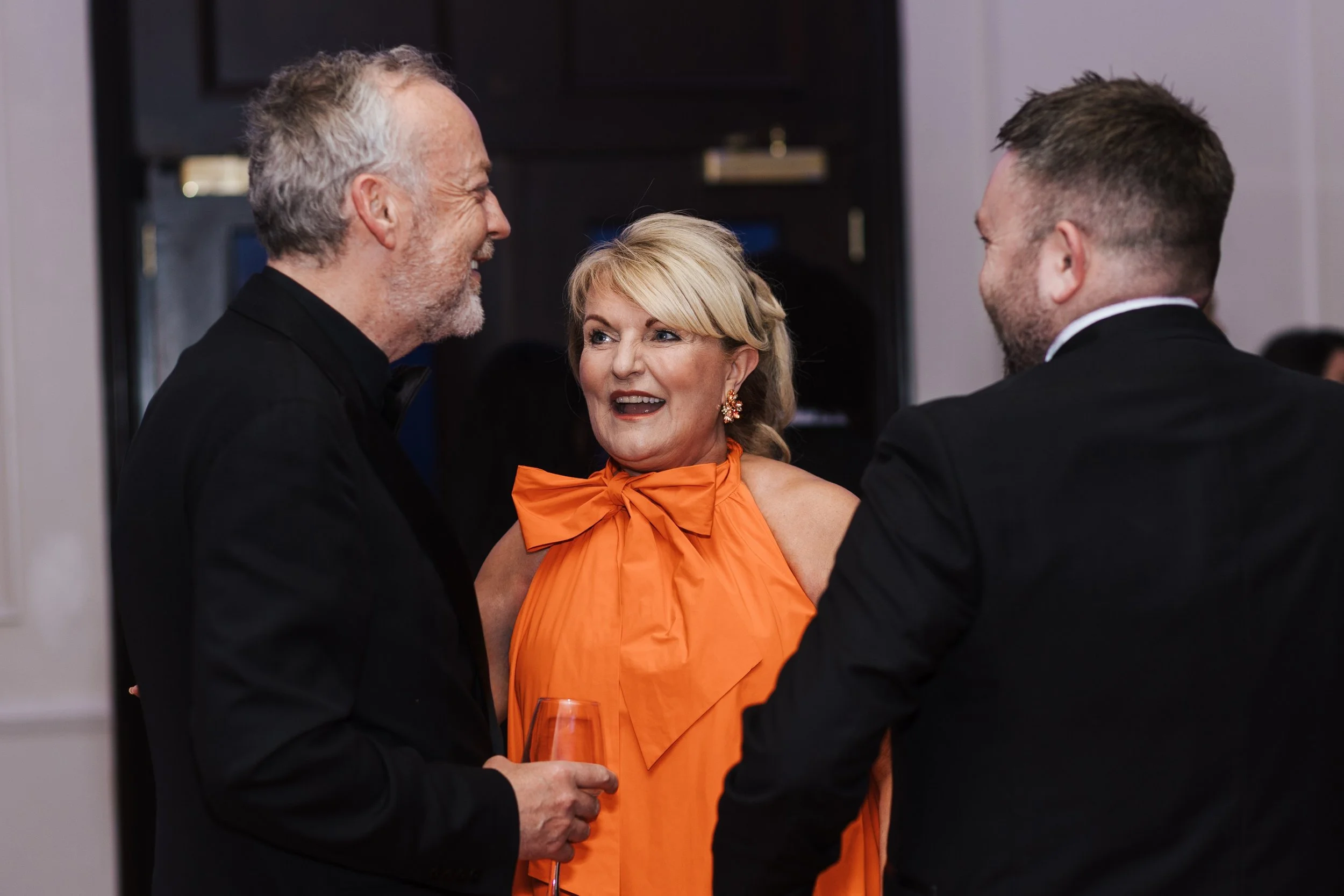 Three people having a lively conversation at a social event. An older man with gray hair and a beard is smiling and holding a glass of wine. A woman in the center, wearing an orange dress with a bow, is engaged in the discussion. A younger man with s