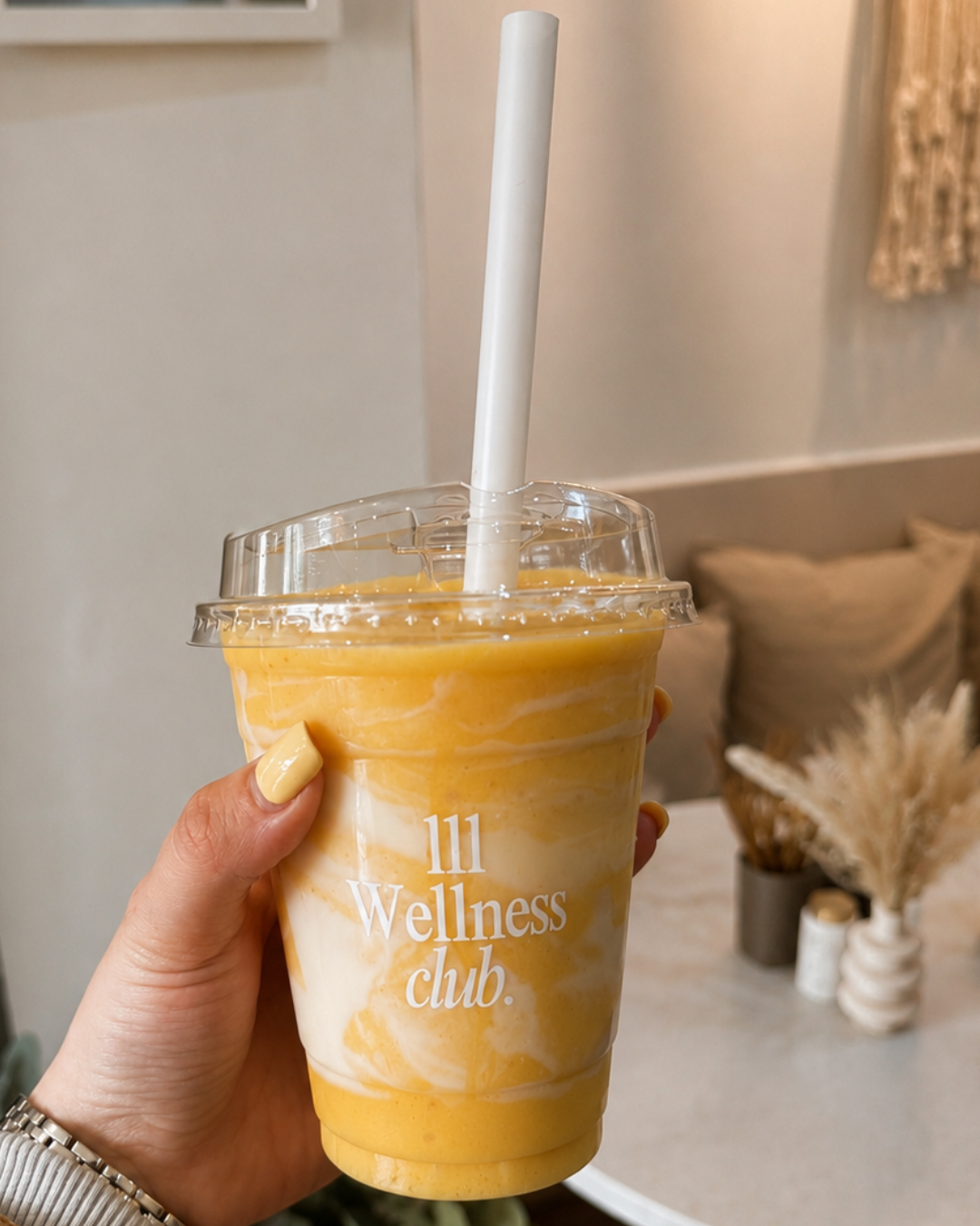 A hand holding a clear plastic cup with a white and orange swirled smoothie, with a white straw. The cup has '111 Wellness club' written on it. In the background, there is a beige wall, a table with a small vase of dried flowers, and some pillows.