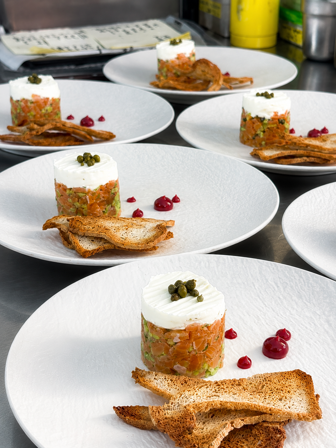A plated starter course of tuna tartare with a round whipped cheese topped with capers, served with triangular toast and a few red wine sauce droplets on a textured white plate.
