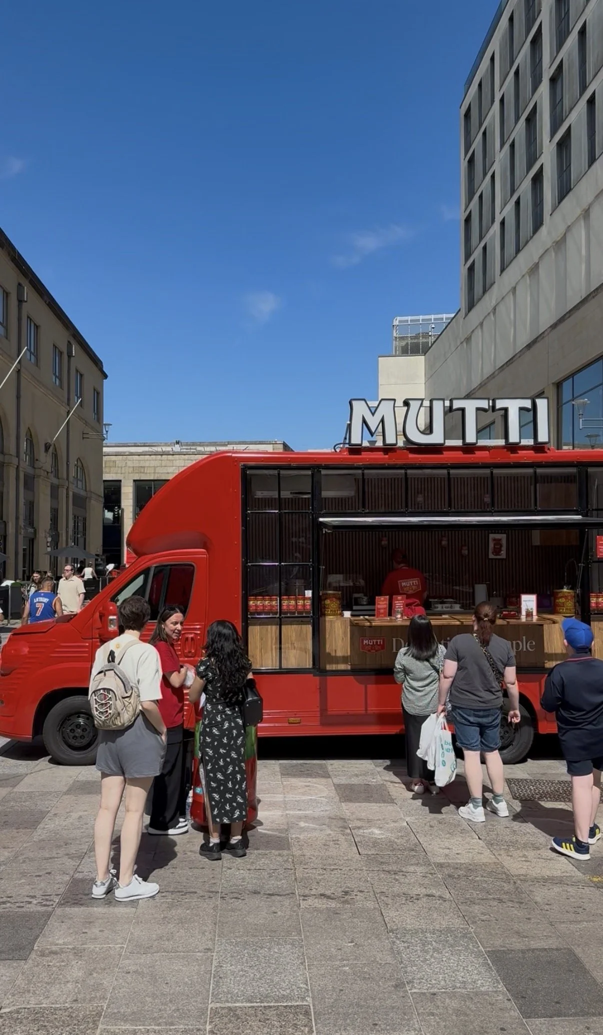 A red food truck with the word 'MUTTI' on the sign on top, serving people in an outdoor urban area with tall buildings and a clear blue sky.