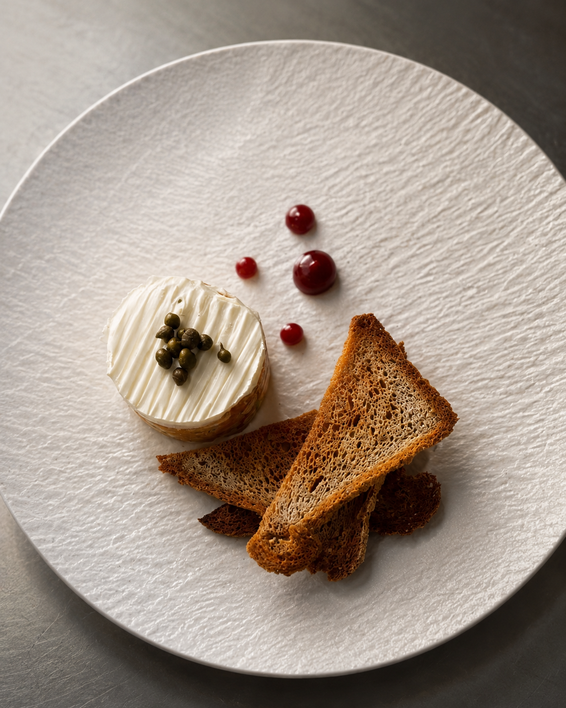 A plated starter course of tuna tartare with a round whipped cheese topped with capers, served with triangular toast and a few red wine sauce droplets on a textured white plate.