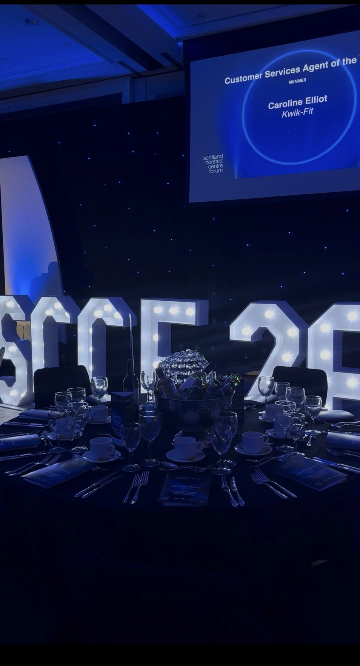 Award ceremony reception table with illuminated large letters spelling 'SCCF' and '26', set with glassware, plates, and utensils, with a dark background, large screen displaying award details, and a dark starry backdrop.